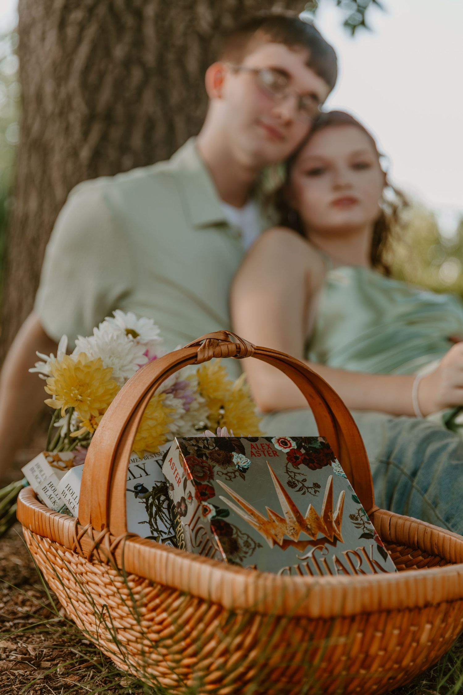 A romantic countryside picnic scene with a woven basket containing flowers and a couple relaxing against a tree trunk.