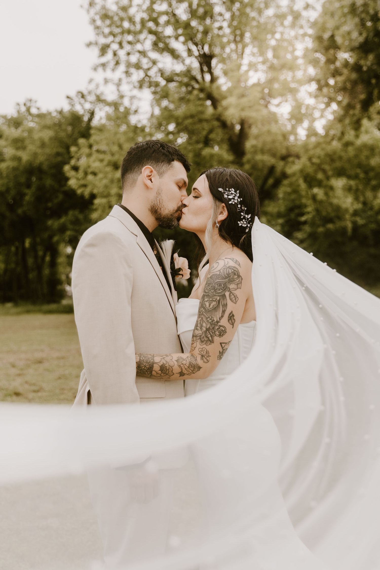 Bride and groom sharing a romantic kiss outdoors, with veil flowing, groom wearing tattoos and beige suit.