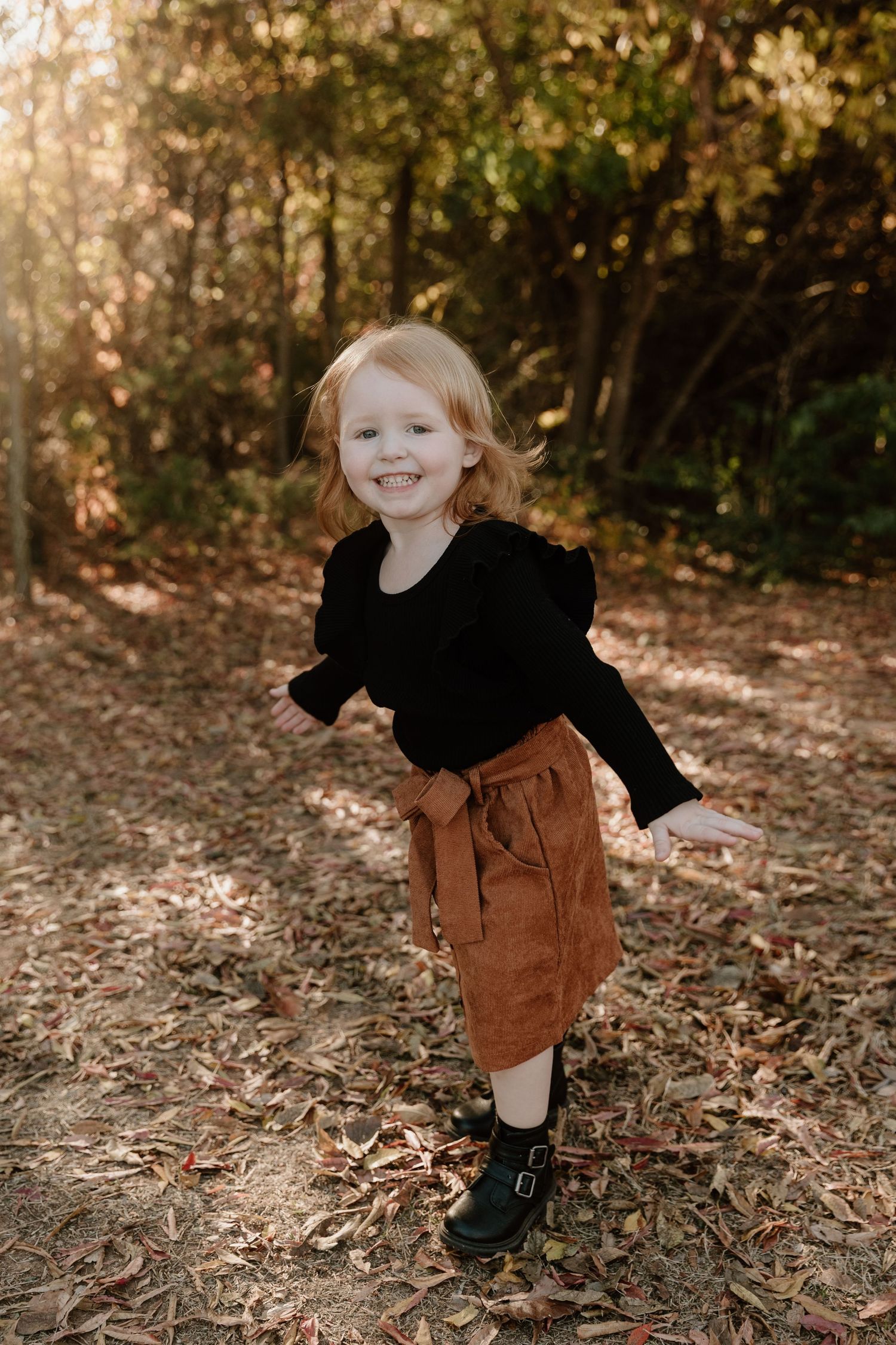 A young child in a black sweater and brown skirt poses playfully on fallen leaves in a sunlit forest setting.