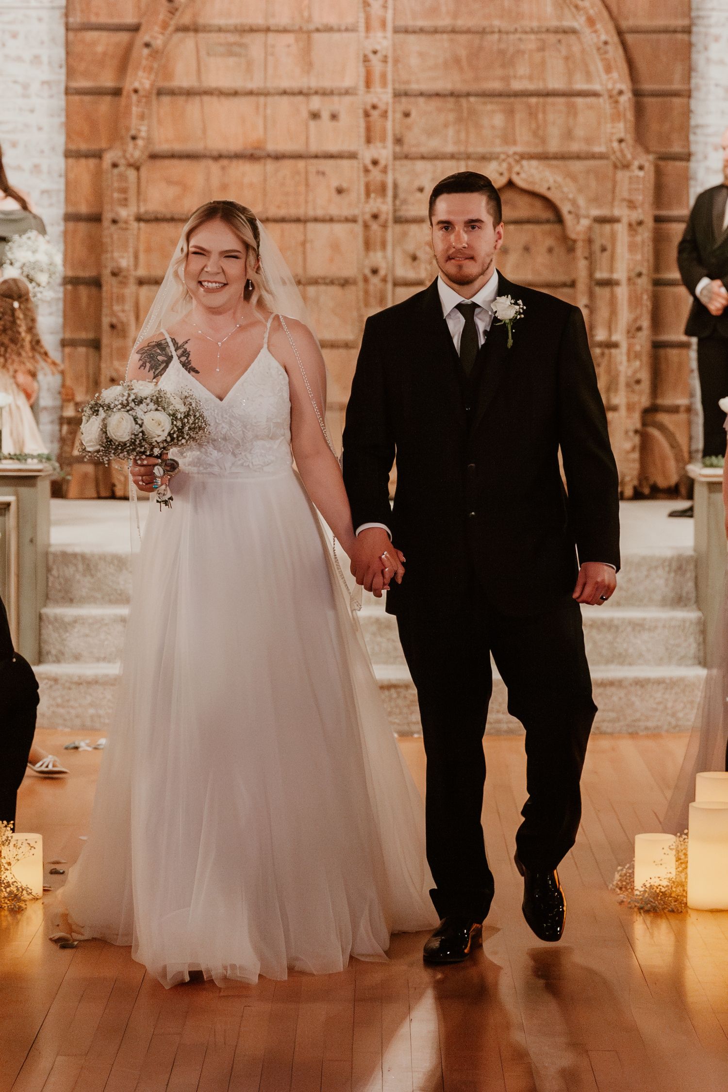 A couple walks down a candlelit aisle in a stunning indoor wedding ceremony with warm brick walls.