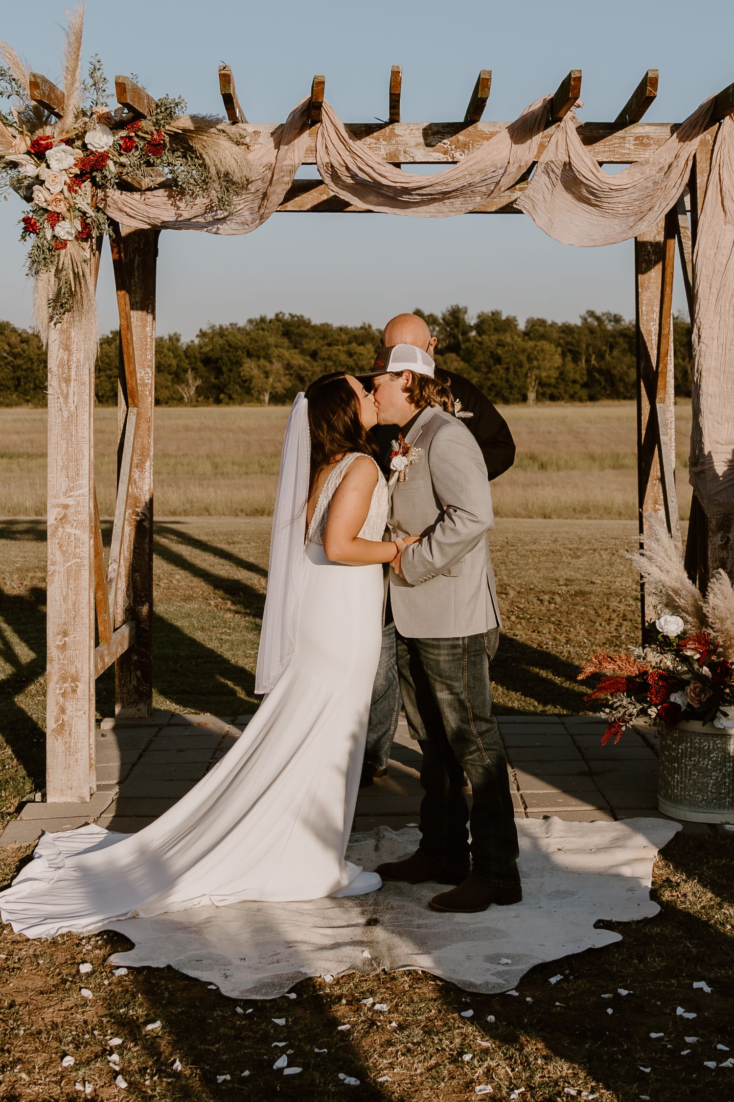 Romantic wedding couple shares intimate moments during sunset ceremony under rustic wooden arch decorated with flowers.