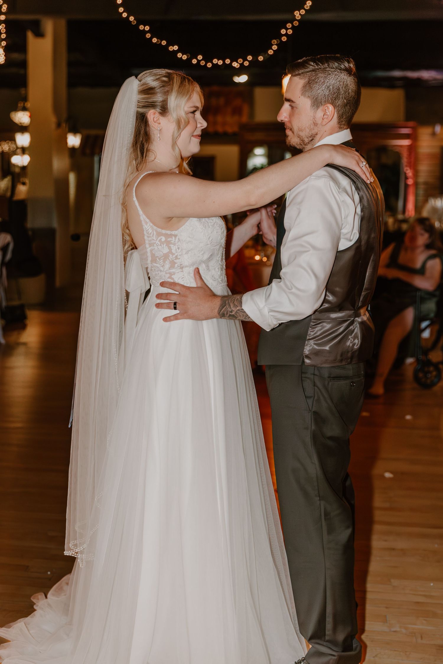 A newlywed couple shares their first dance at a wedding reception under string lights on a wooden dance floor.