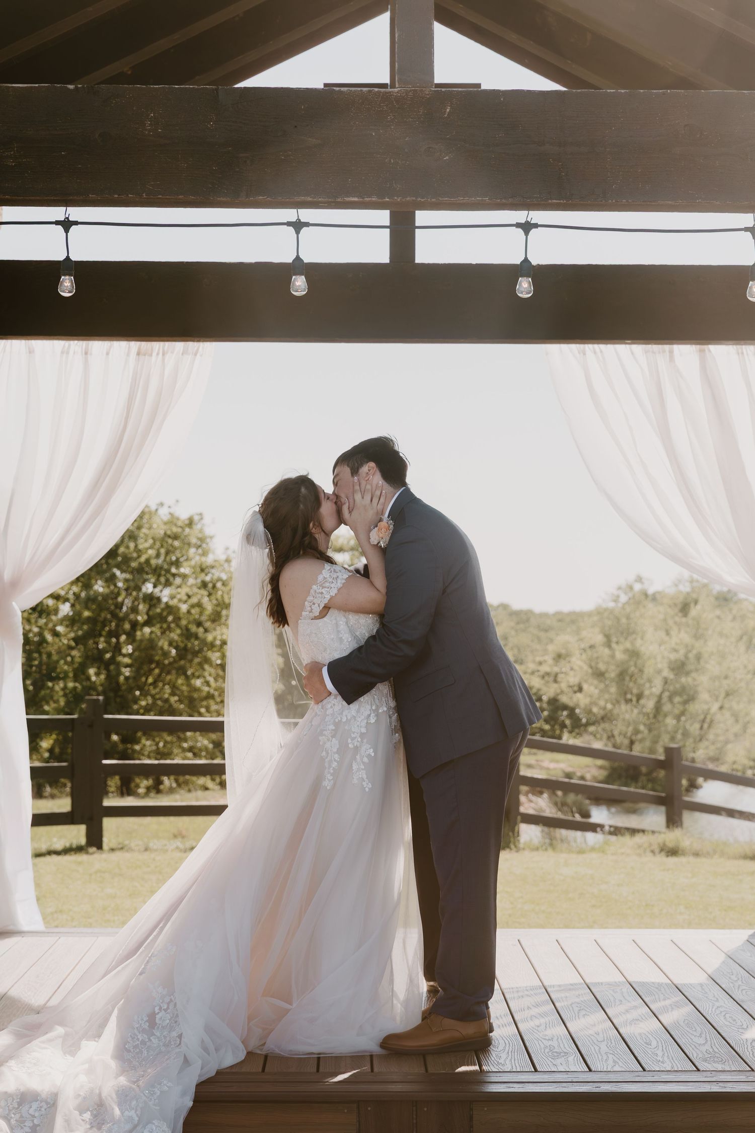 Bride and groom share romantic first kiss under rustic wooden pavilion with sheer curtains outdoors.