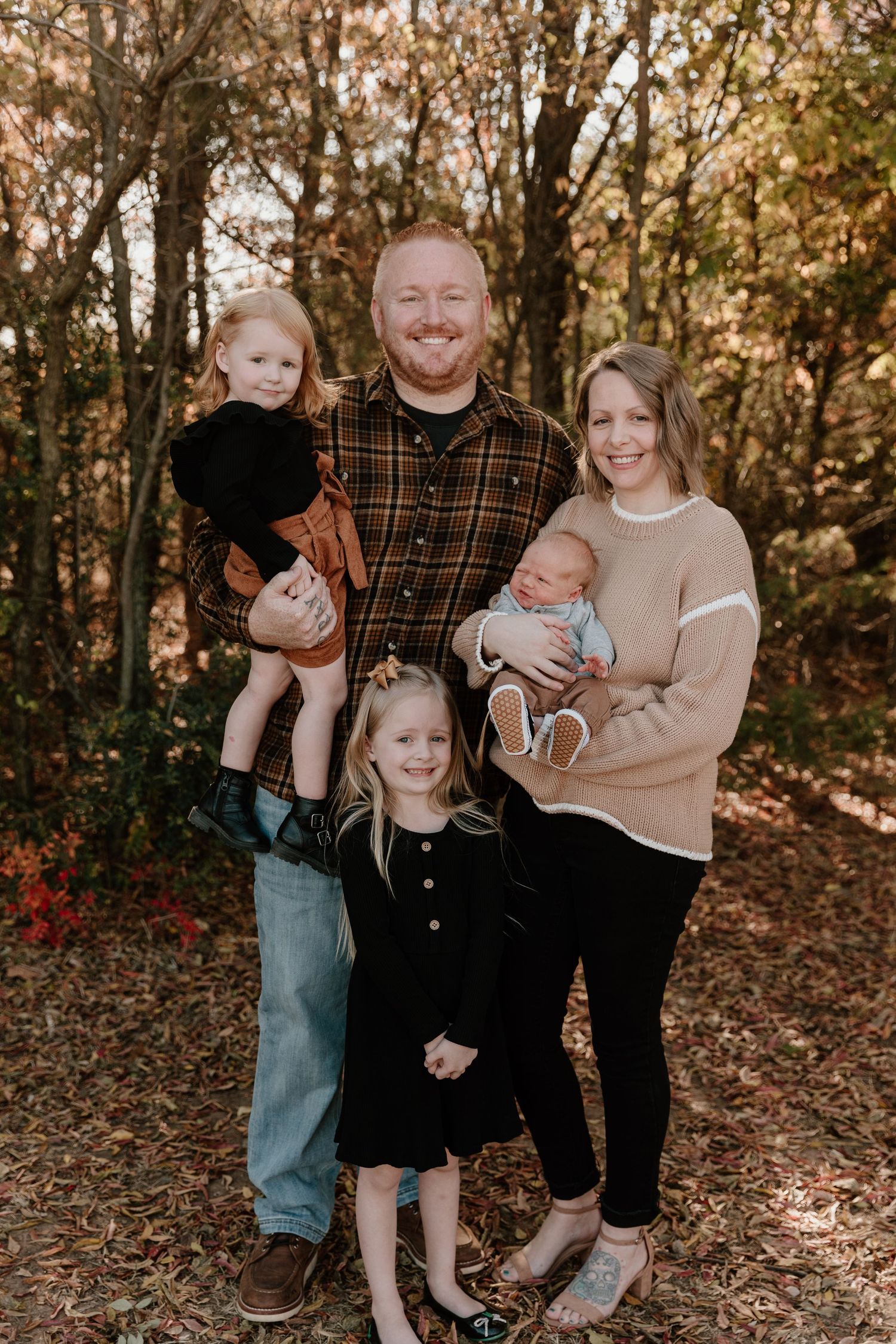 A family poses together for a casual autumn photoshoot in a wooded setting with fallen leaves.