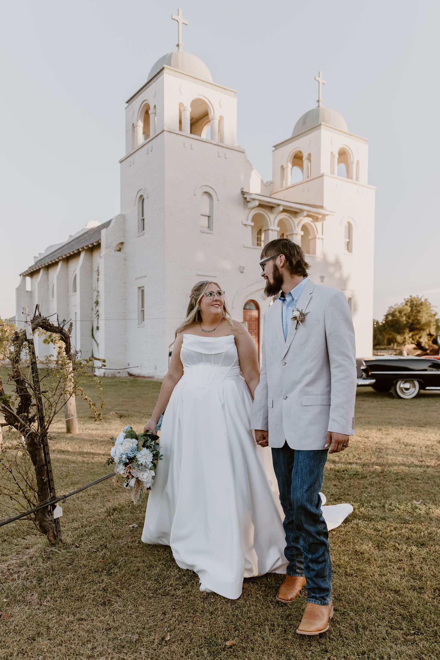 A couple poses for wedding photos in front of a white Spanish mission-style church with multiple bell towers.