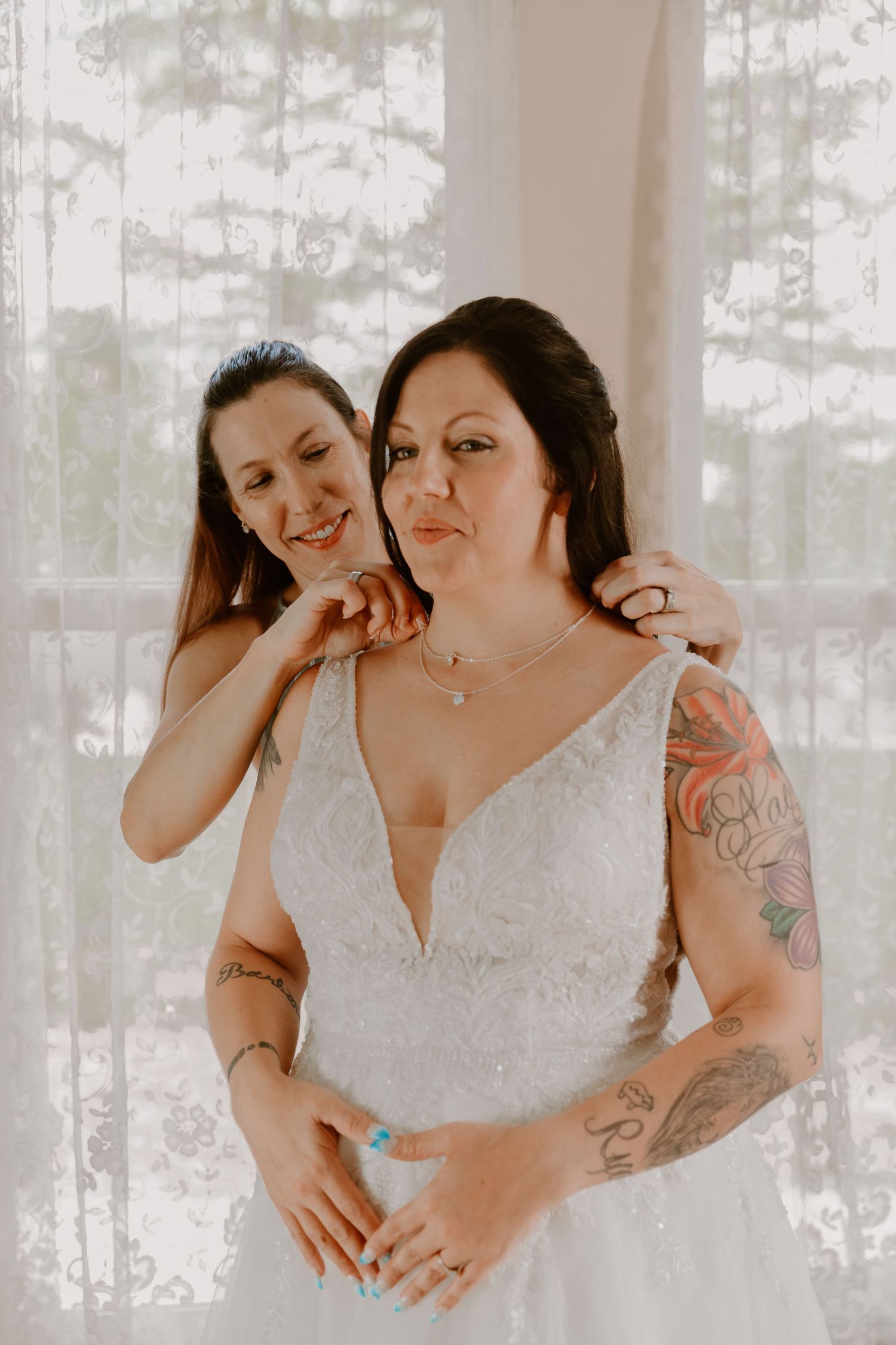 An intimate pre-wedding moment between two people in white lace dress with visible tattoos against sheer curtains.
