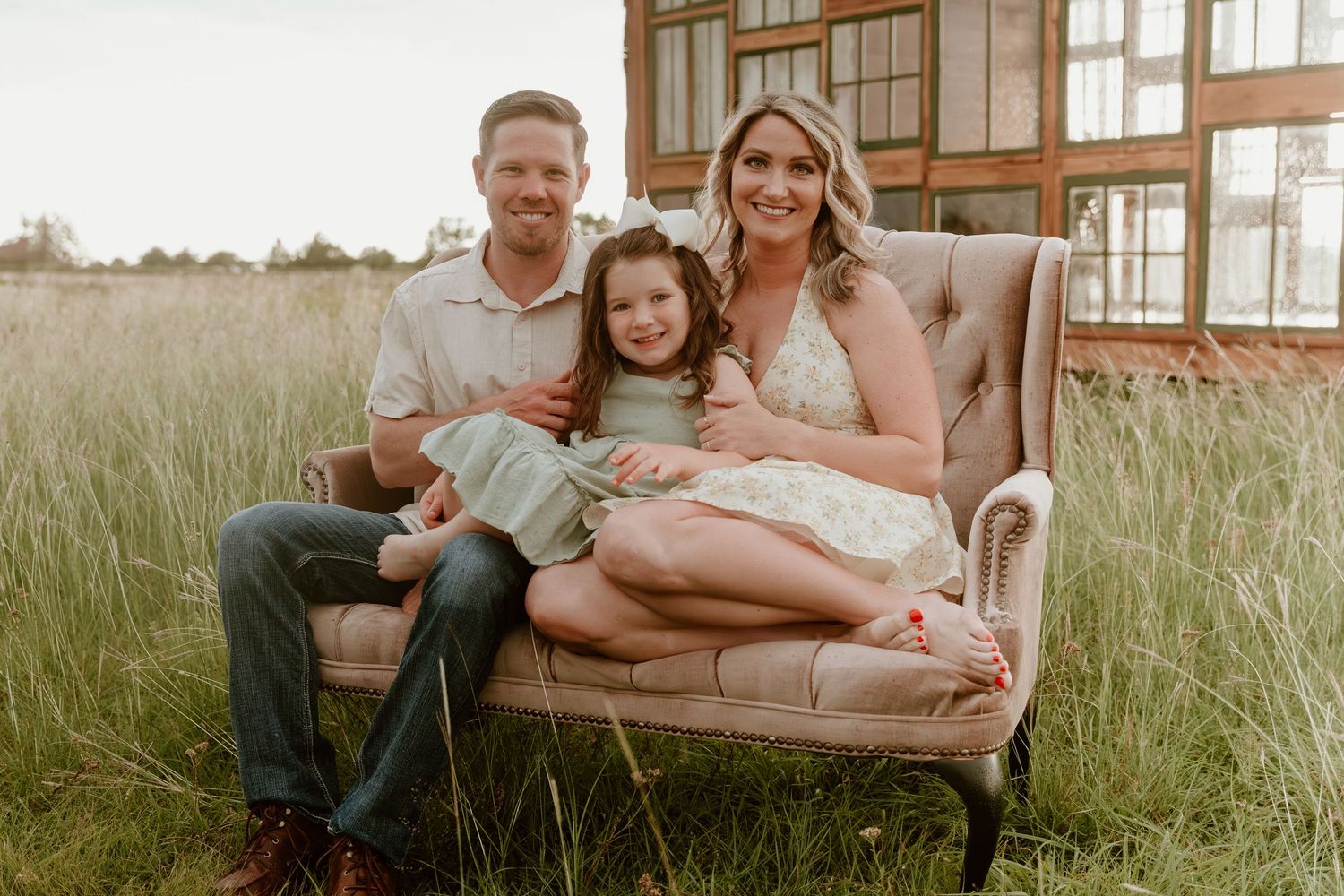 Family sits together on a vintage beige couch in a sunny meadow with tall grass and golden light.
