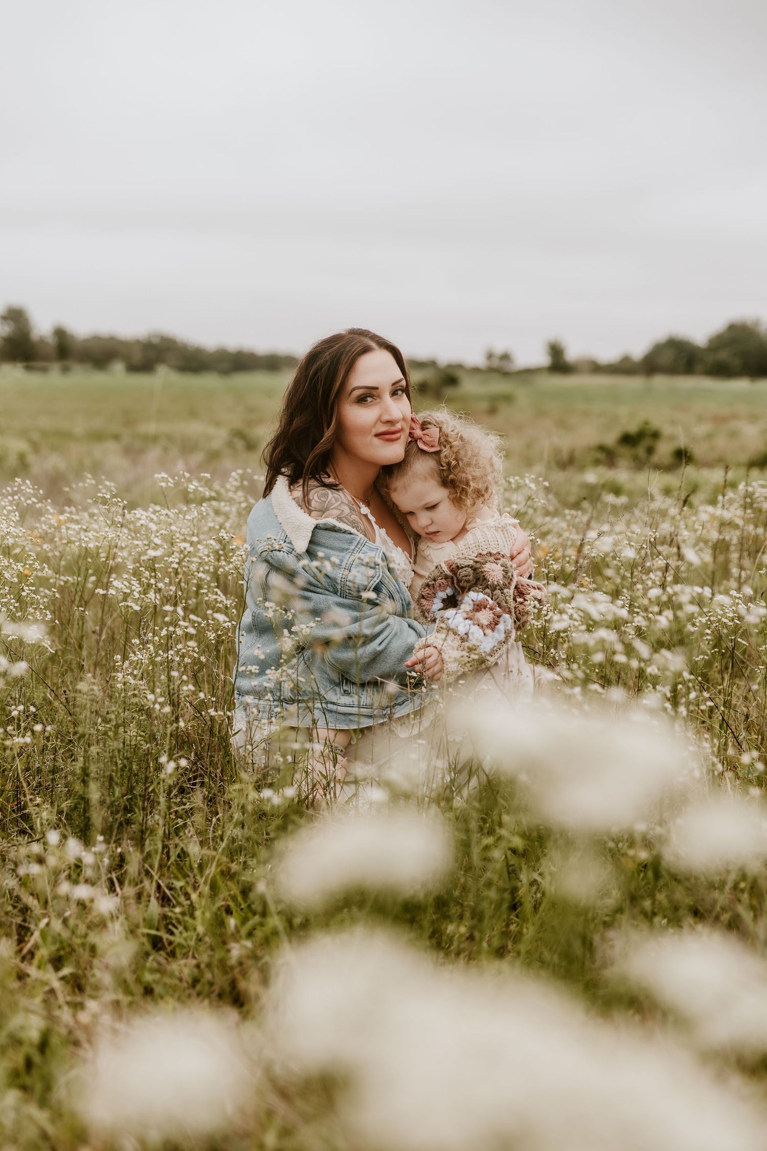 A mother and young child sit together in a blooming wildflower field, sharing a tender embrace on an overcast day.