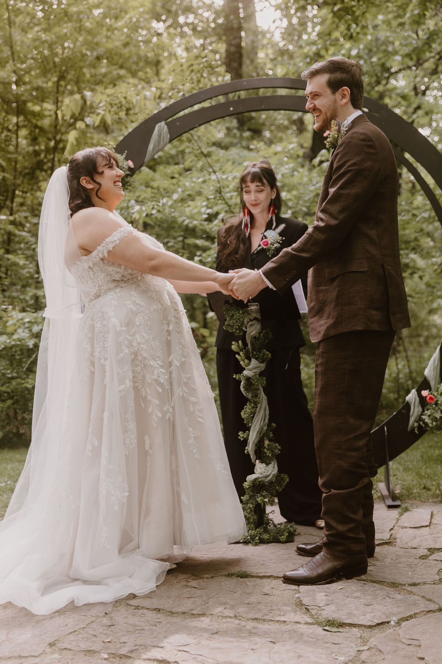 Bride and groom hold hands smiling at outdoor wedding ceremony under circular arch with greenery in wooded setting.