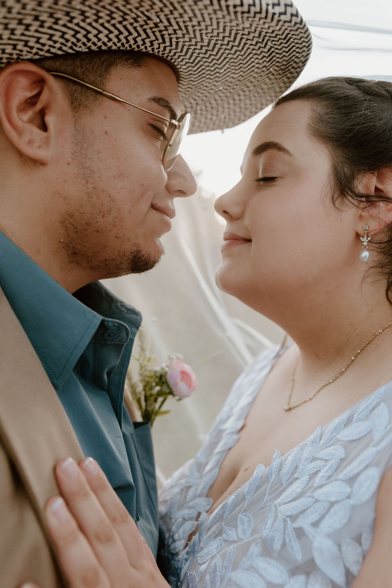 Intimate close-up of bride and groom almost kissing under wedding veil, groom in teal shirt.