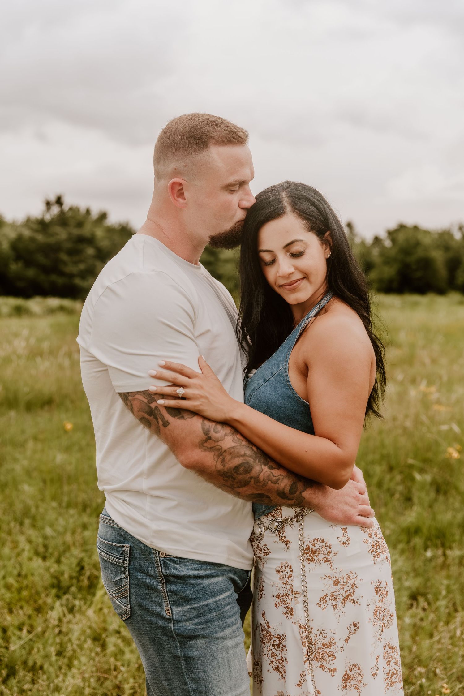 A couple embraces in a green field, the man kissing her head, both wearing casual summer outfits with tattoos visible.