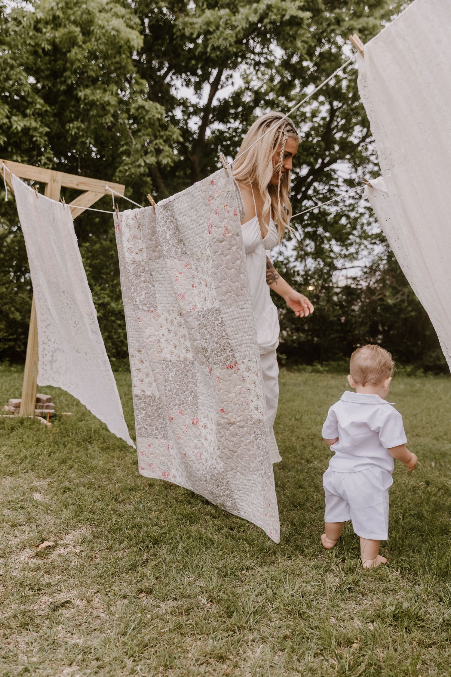 Blonde woman hanging a patchwork quilt on a clothesline outdoors while a toddler in white stands nearby on green grass.