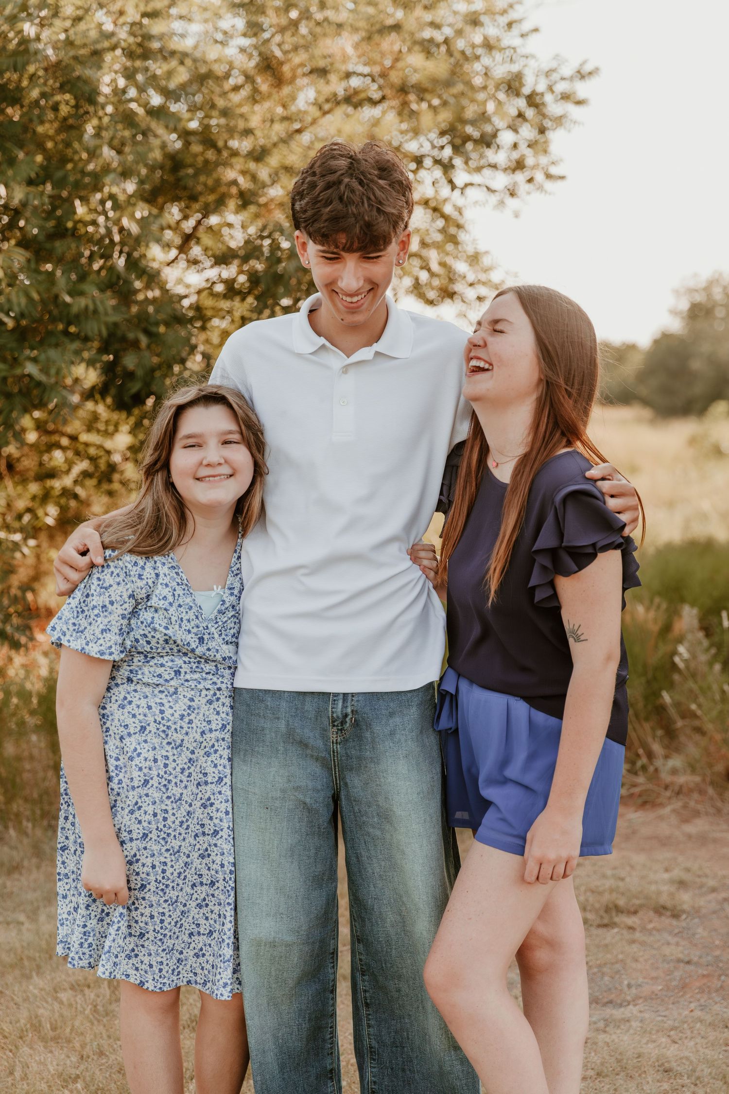 Three people stand together laughing and smiling outdoors in casual clothing on a sunny day.