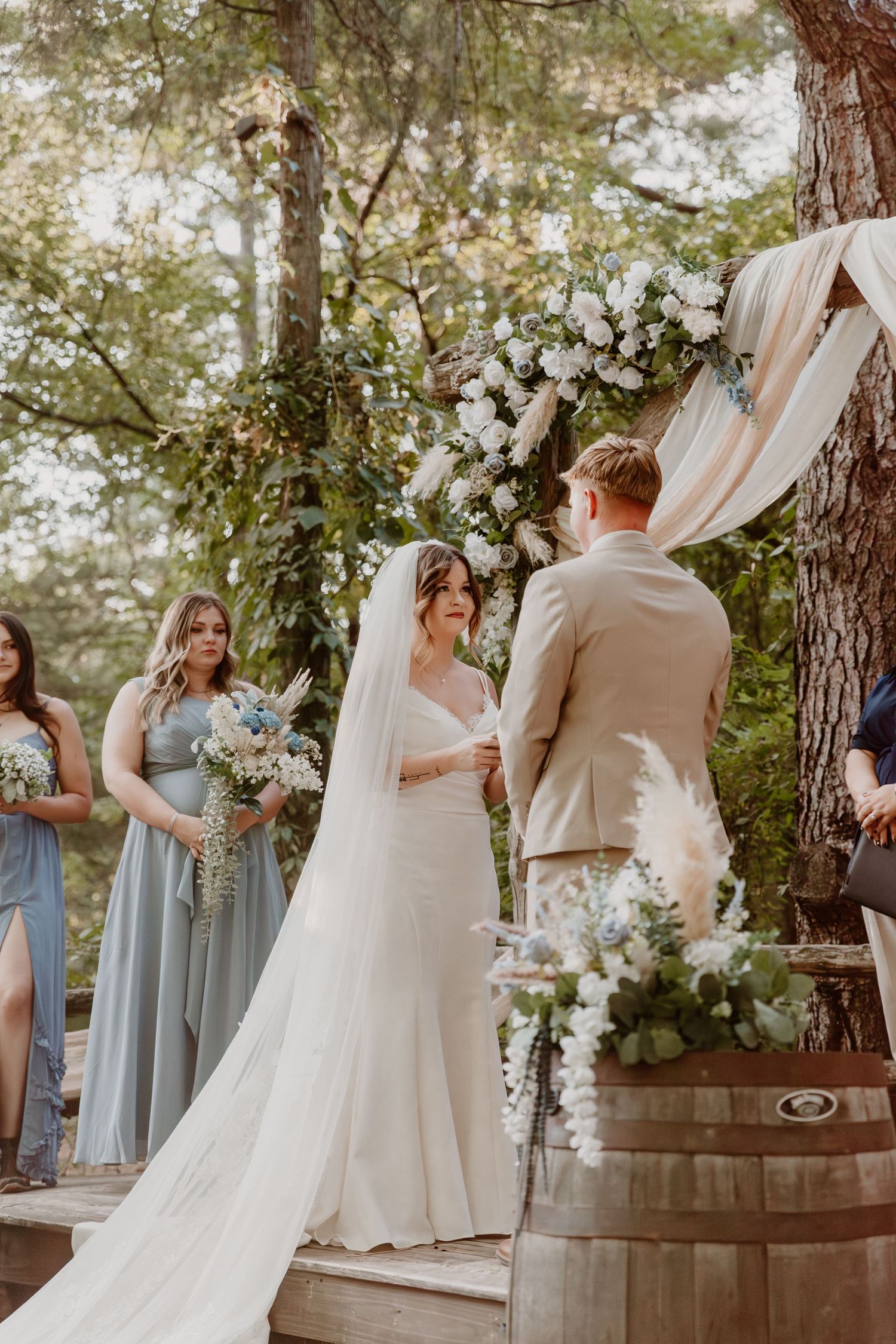 A wedding party in pale blue bridesmaid dresses and a flowing white gown gather outdoors among pine trees.