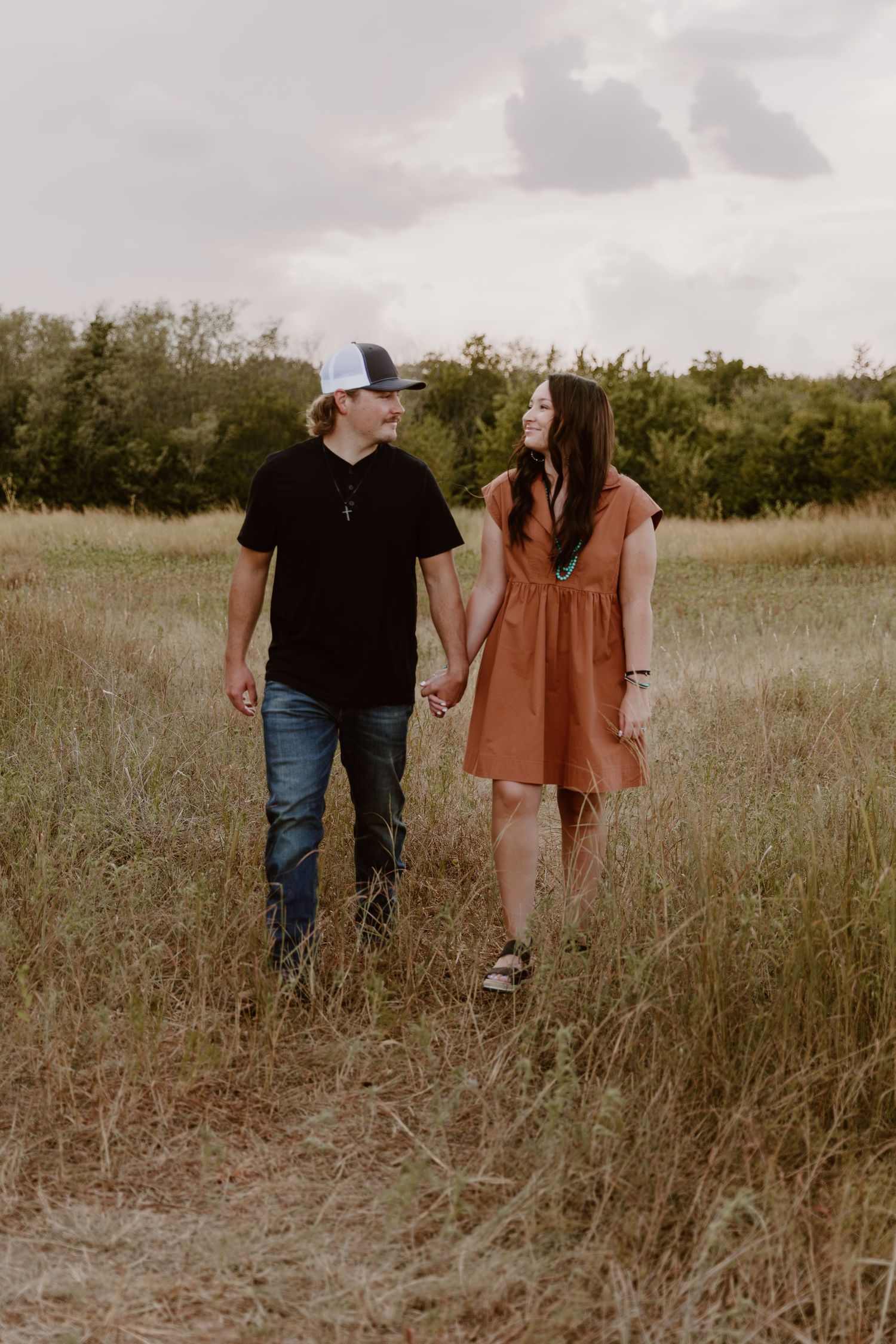 A couple walks hand in hand through a brown grassy field during golden hour.