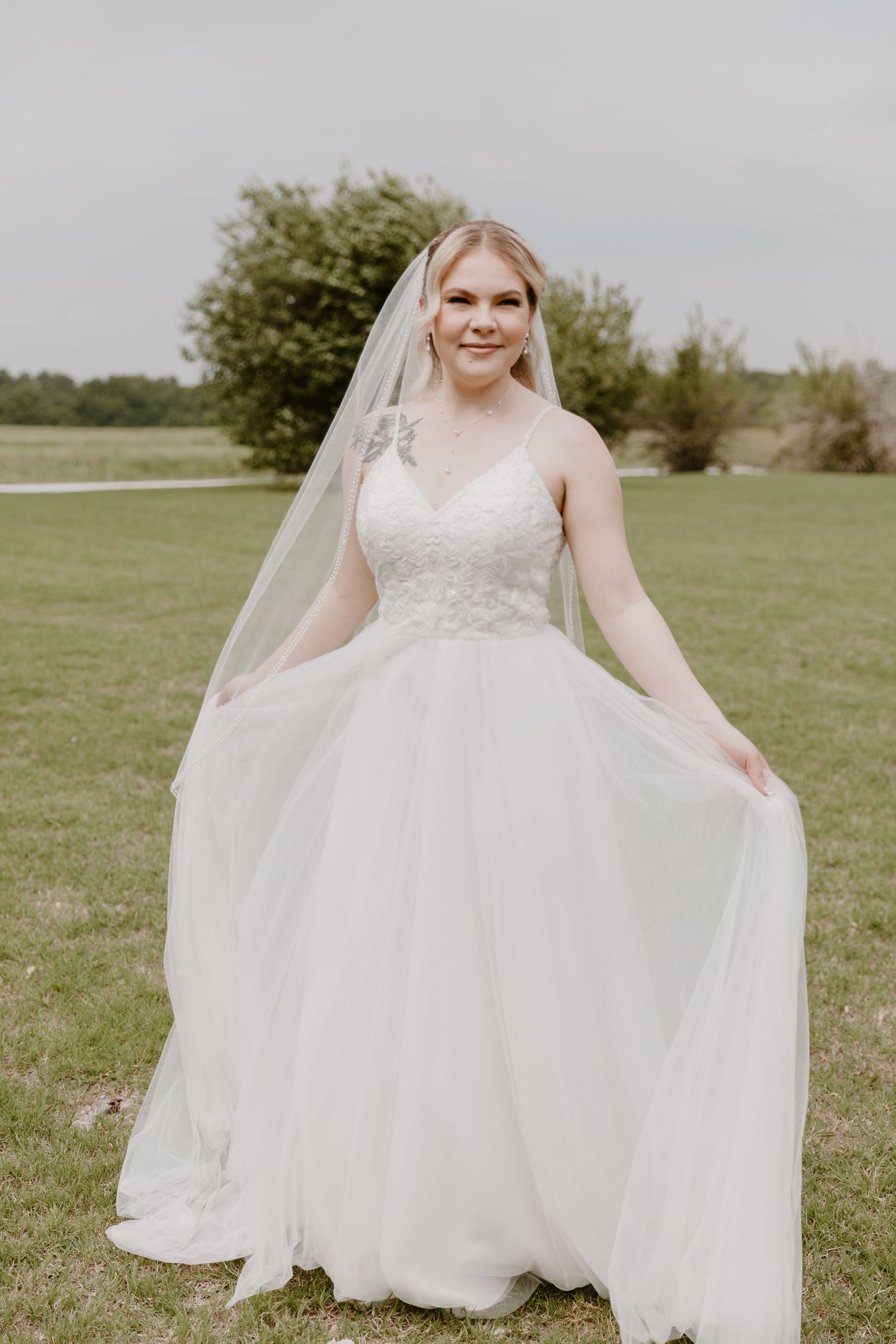 A bride in a flowing white lace and tulle wedding gown stands outdoors on a green lawn holding her dress skirt.
