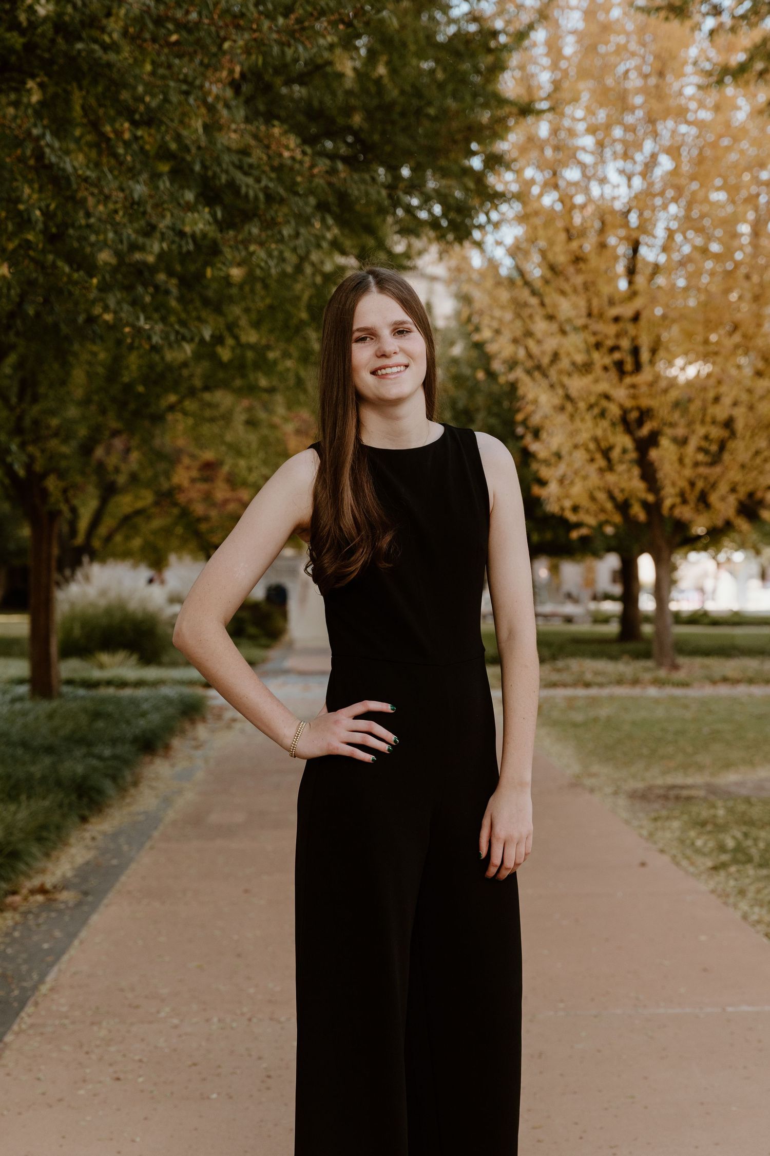 A person in a black sleeveless dress stands on a paved path surrounded by autumn trees with golden foliage.