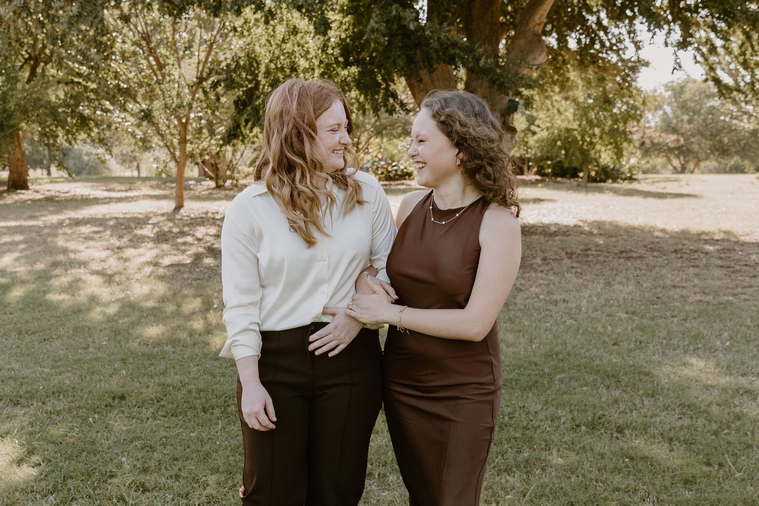 Two people in coordinating brown and white outfits embrace in a sunlit outdoor setting.