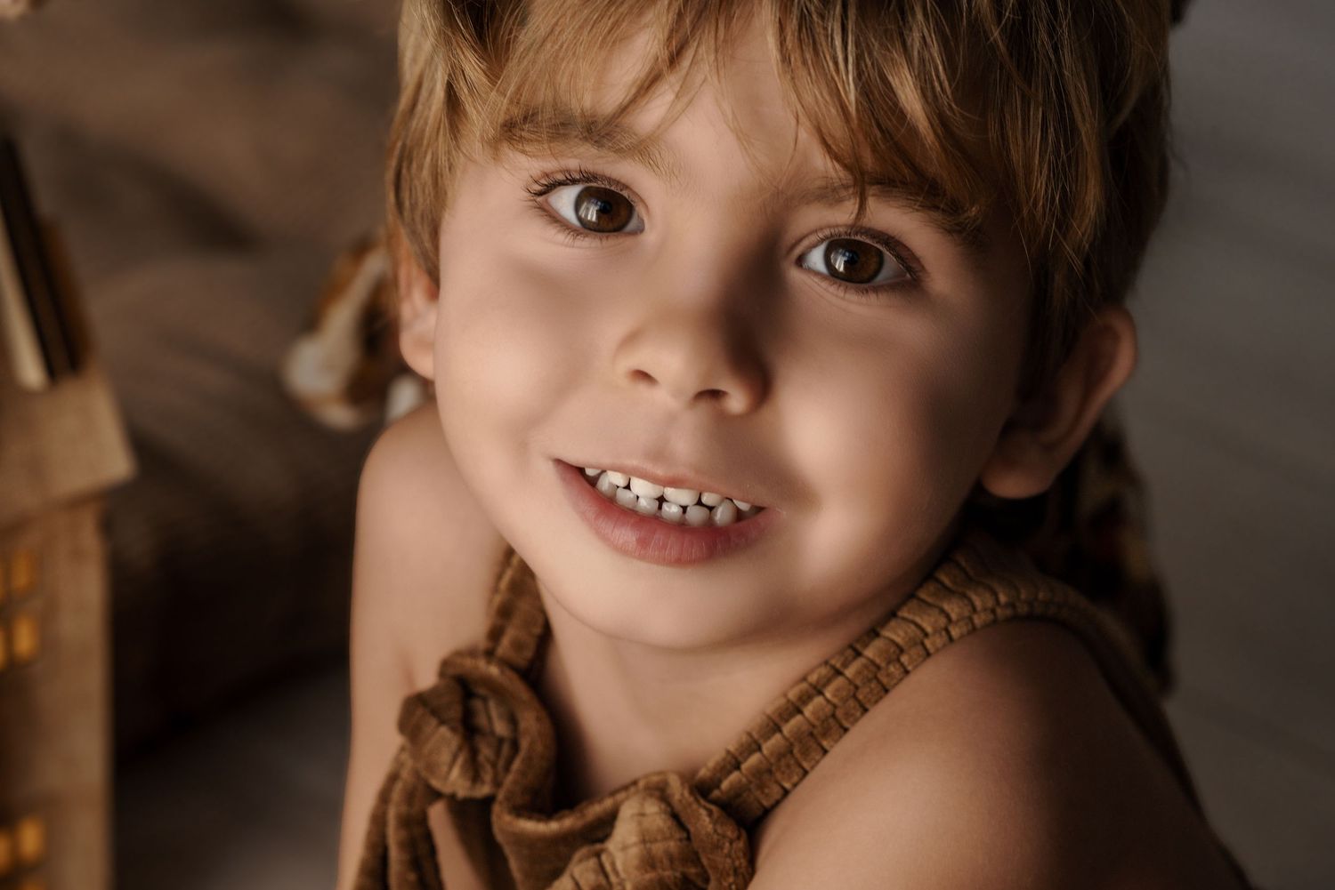 A young child smiles warmly while wearing a brown outfit in warm lighting.