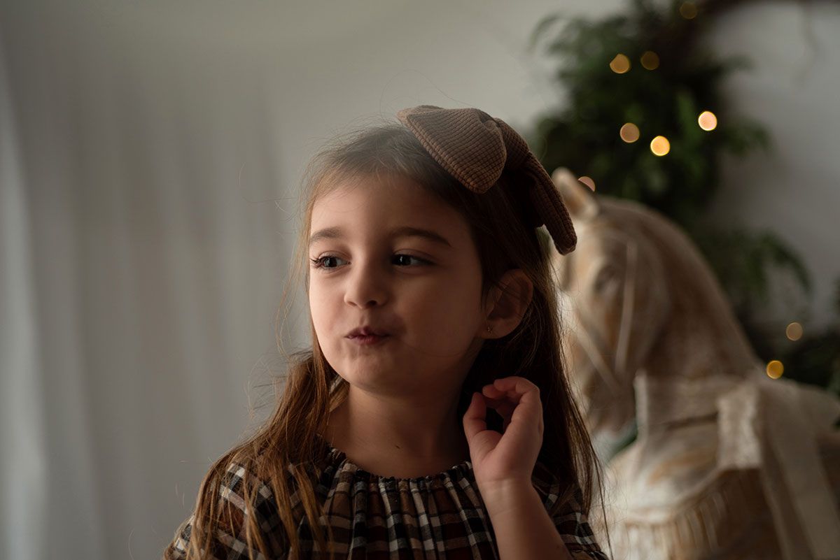 Child in brown patterned dress poses near Christmas tree with soft bokeh lights in background.