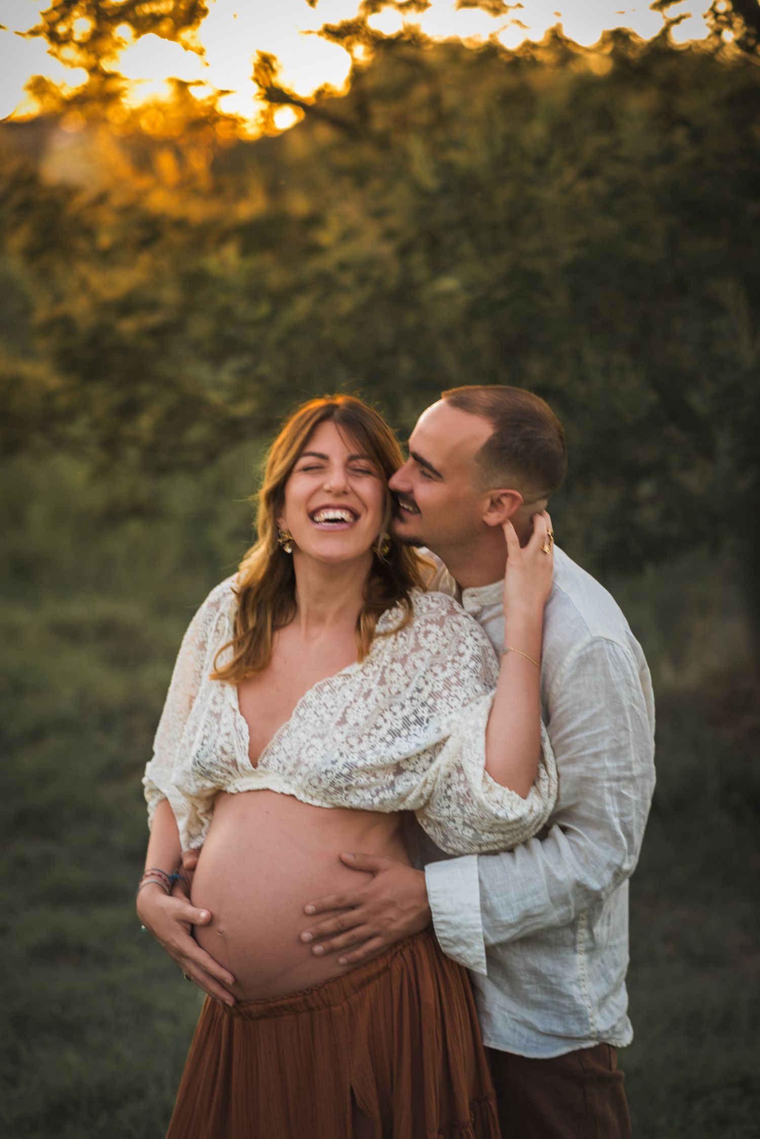 Couple embraces during sunset maternity photoshoot in brown dress and white top against natural backdrop.
