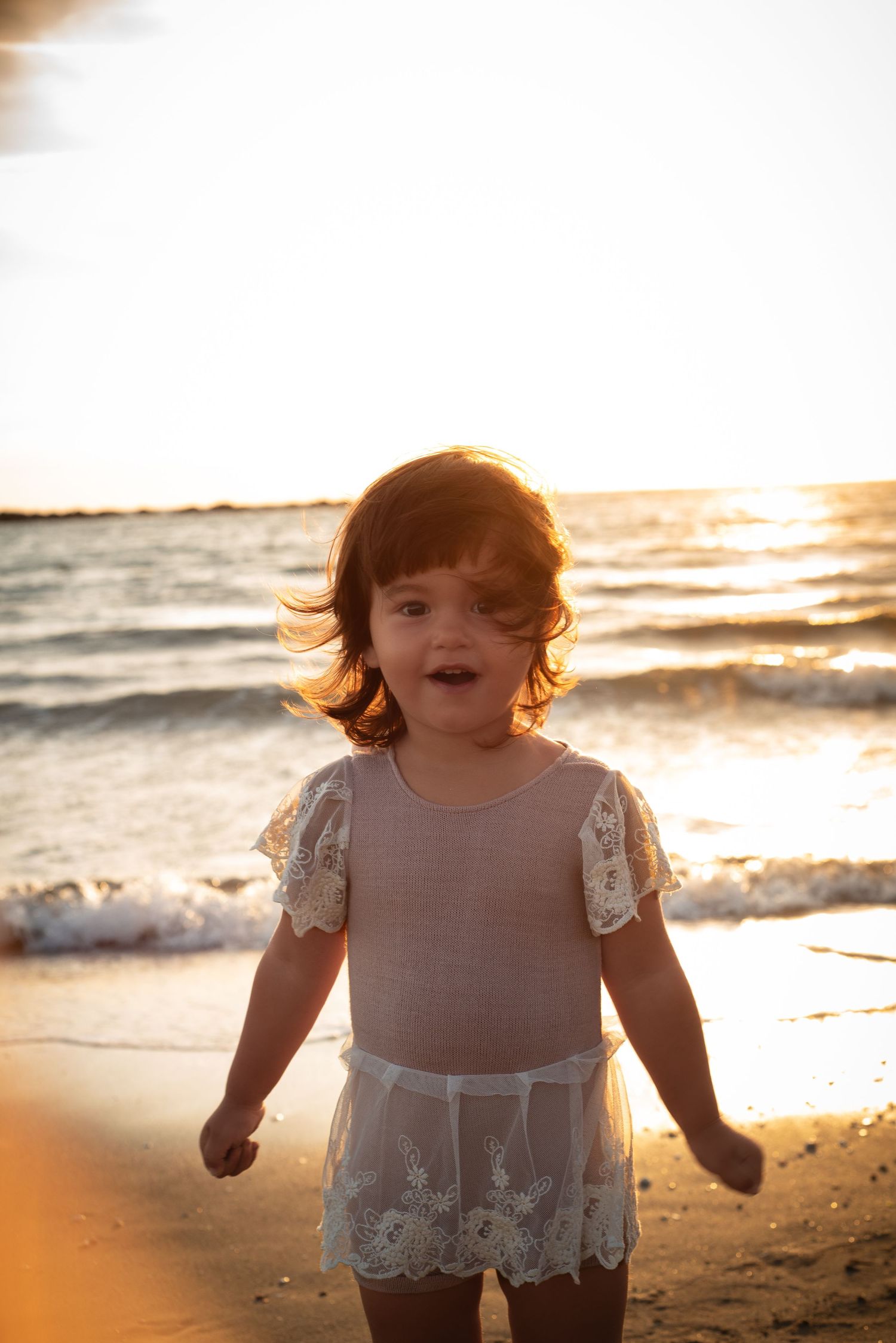 Young child in white lace dress runs joyfully along beach shore at golden sunset.