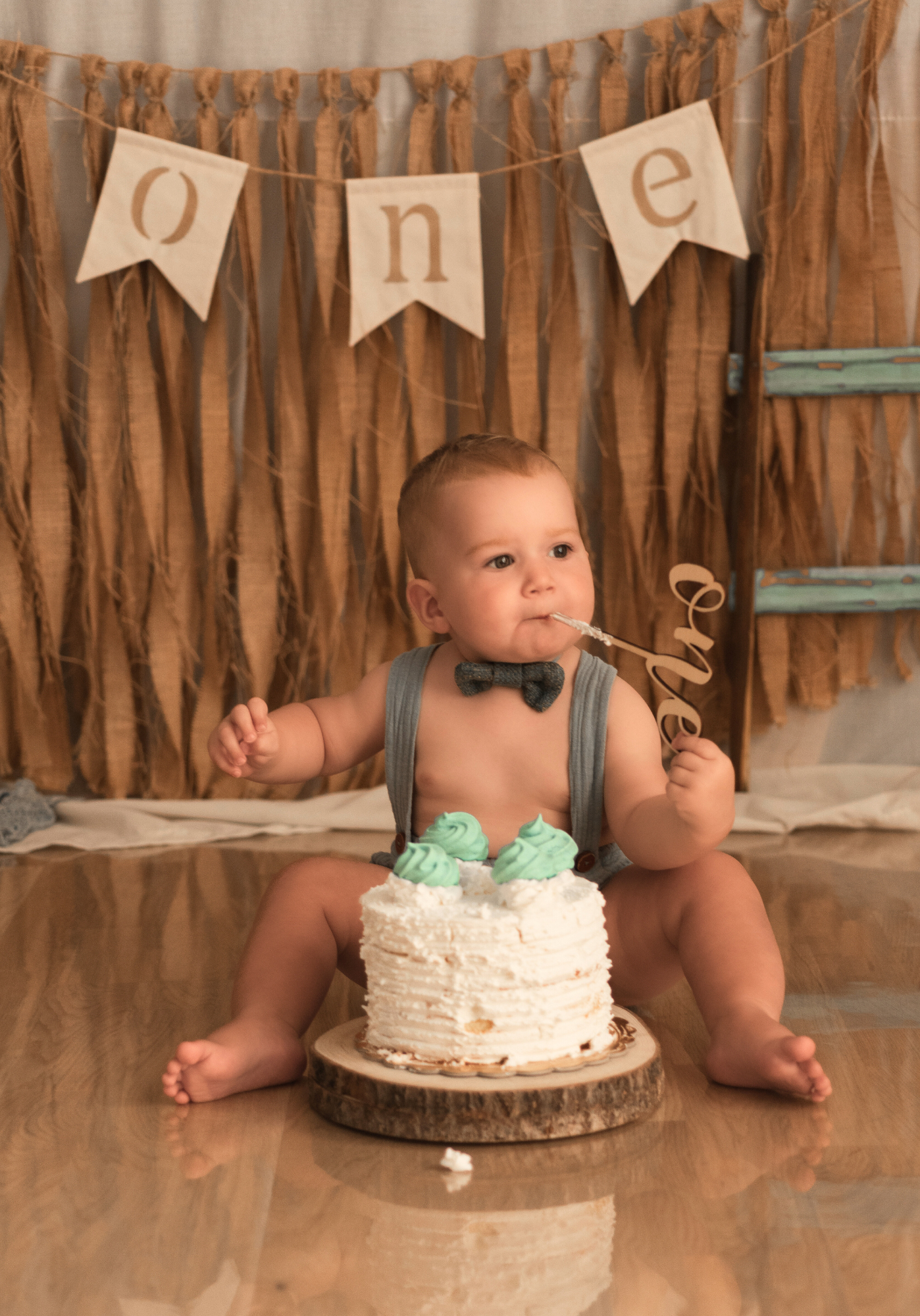 Baby in white diaper sits on floor with birthday cake and rustic burlap backdrop during first birthday photoshoot.