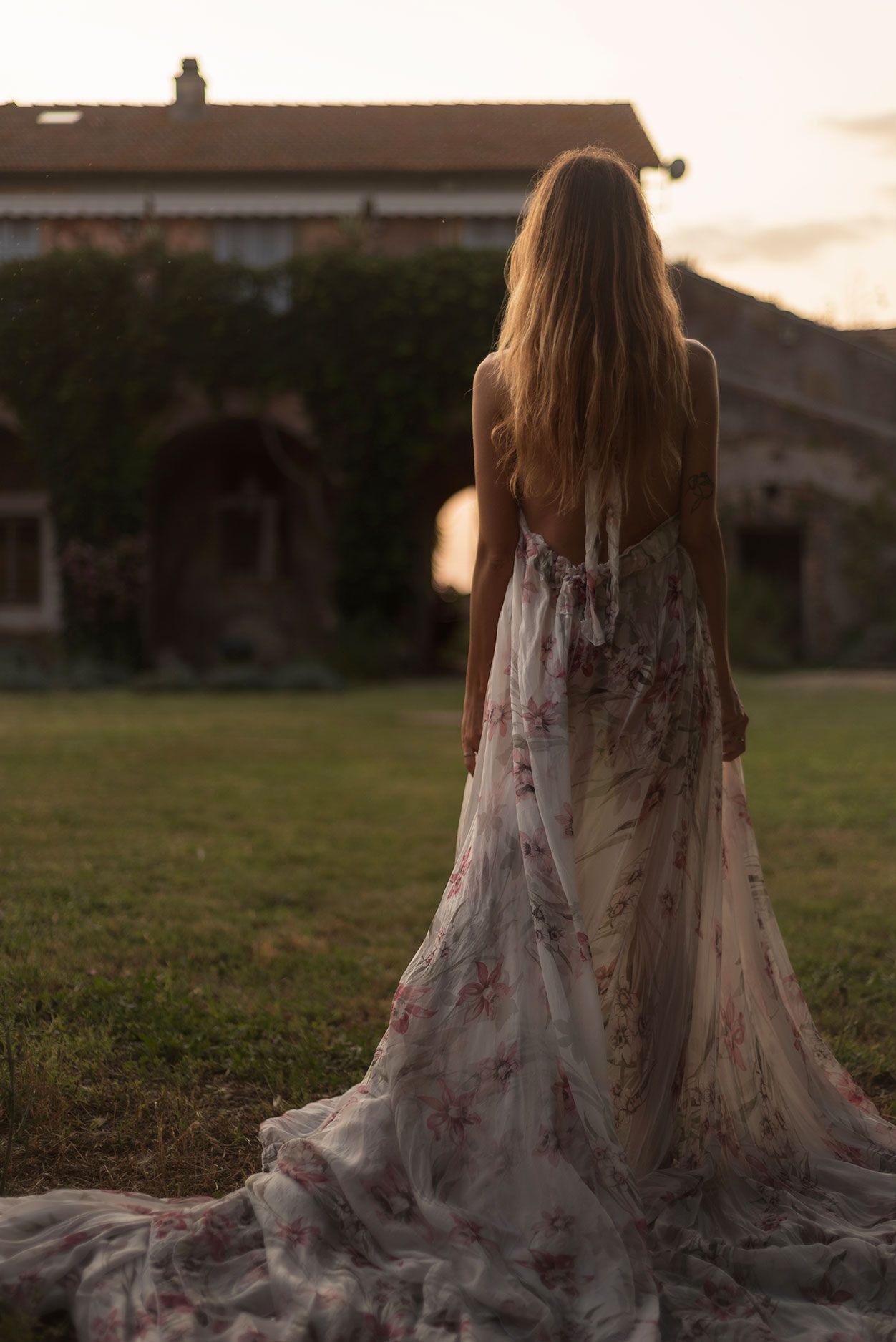 A flowy white floral dress spreads across green grass during golden hour sunset.