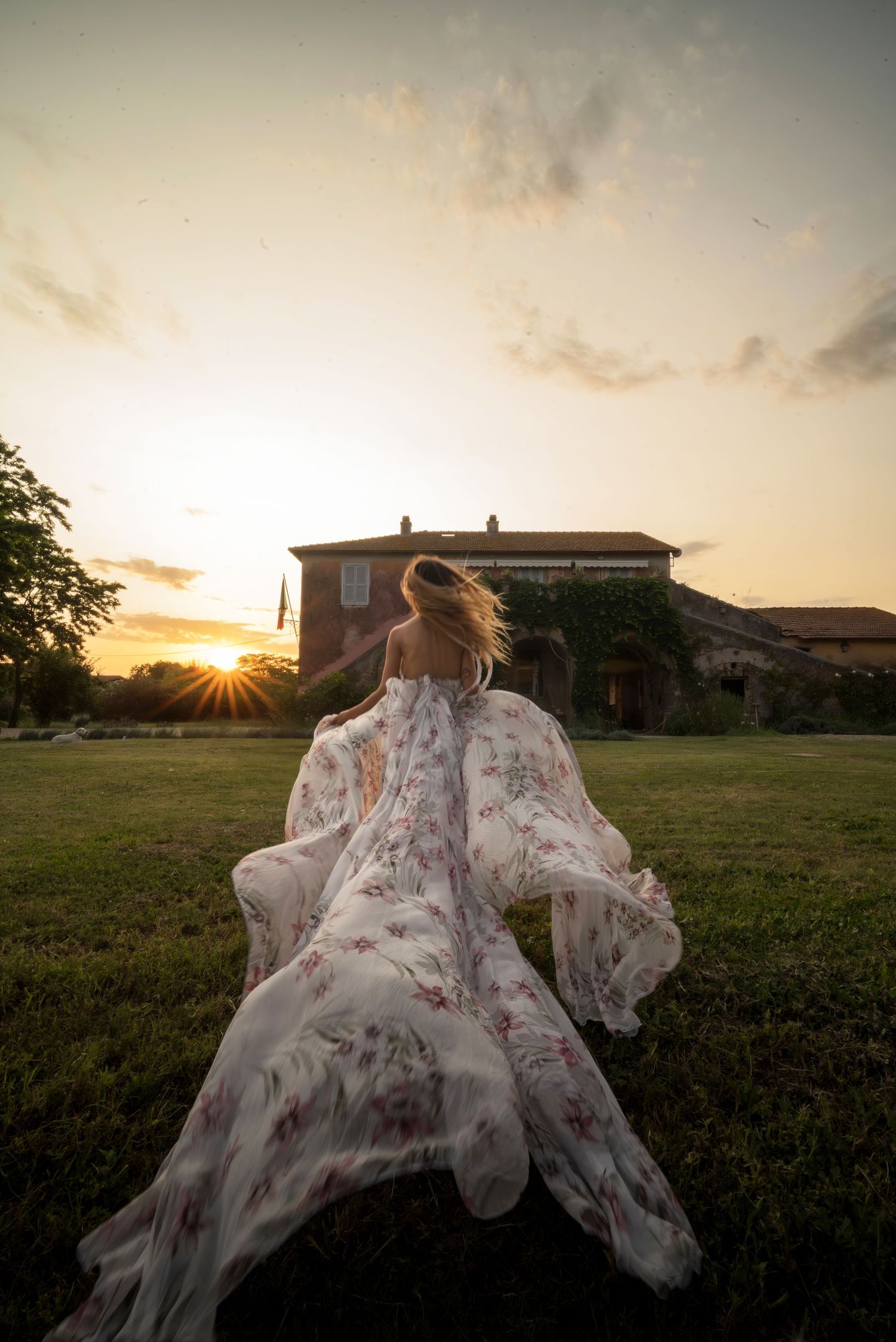 A flowing floral dress billows in the wind as someone runs across a lawn at sunset with a mansion in the background.