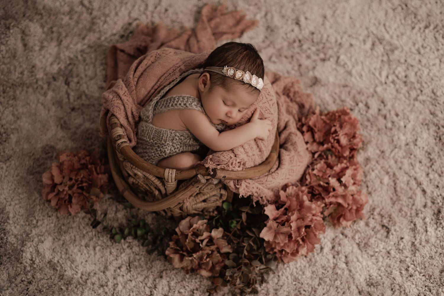 Newborn baby sleeping peacefully in a pink nest surrounded by soft dusty rose flowers on textured backdrop.
