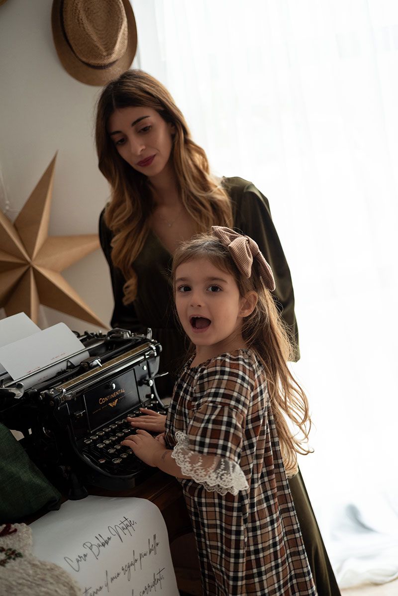 A child in a plaid dress shows excitement while typing on a vintage typewriter next to a decorative gold star on the wall.