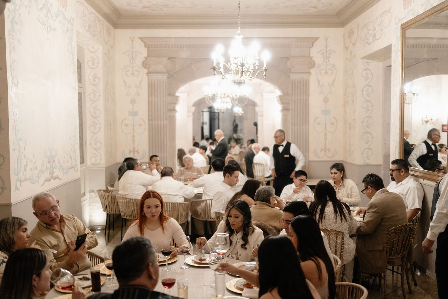 Elegant dining room with crystal chandeliers and white walls filled with guests enjoying dinner at round tables.