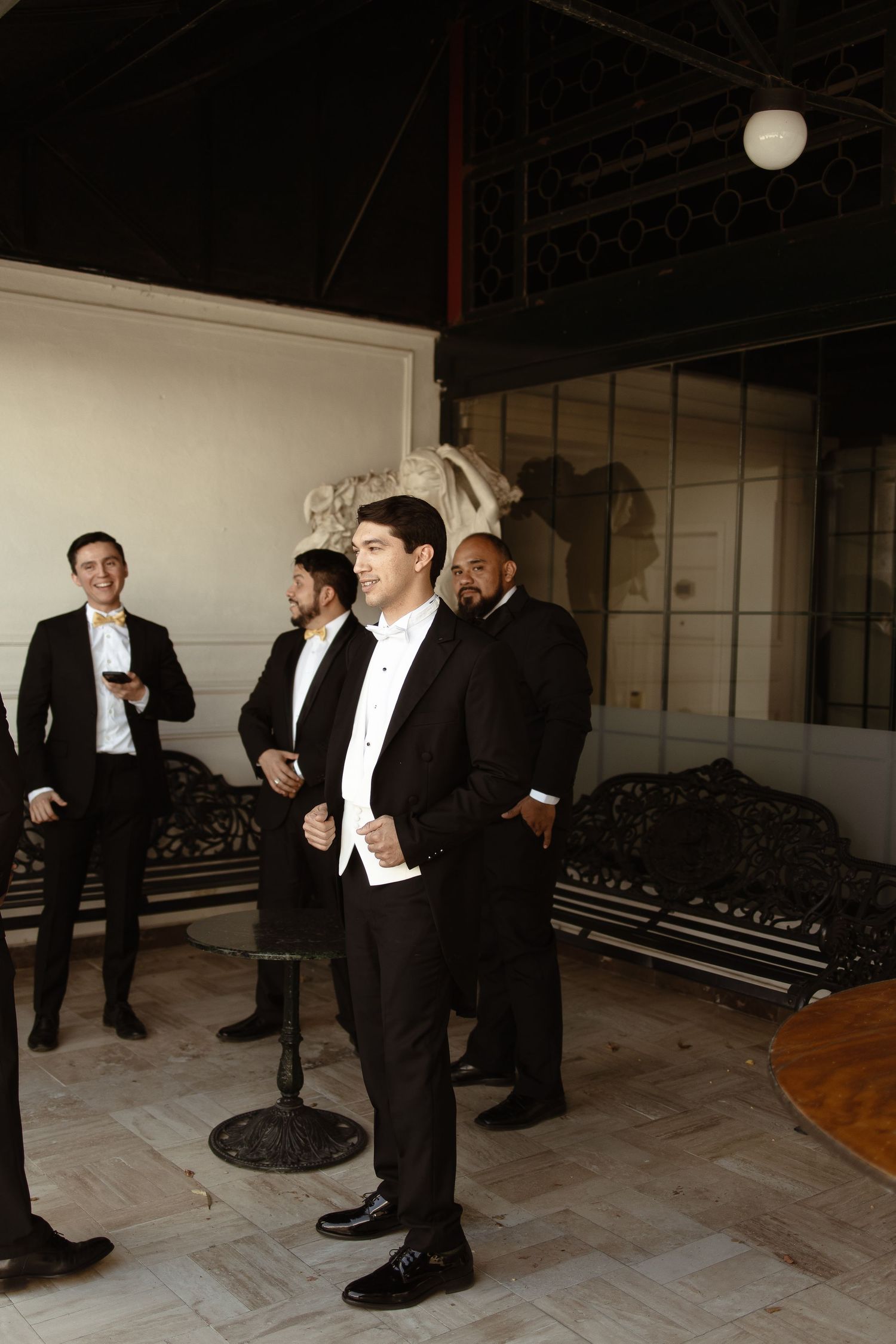 Men in formal black tuxedos and bow ties sit and stand in a backstage dressing area before a performance.