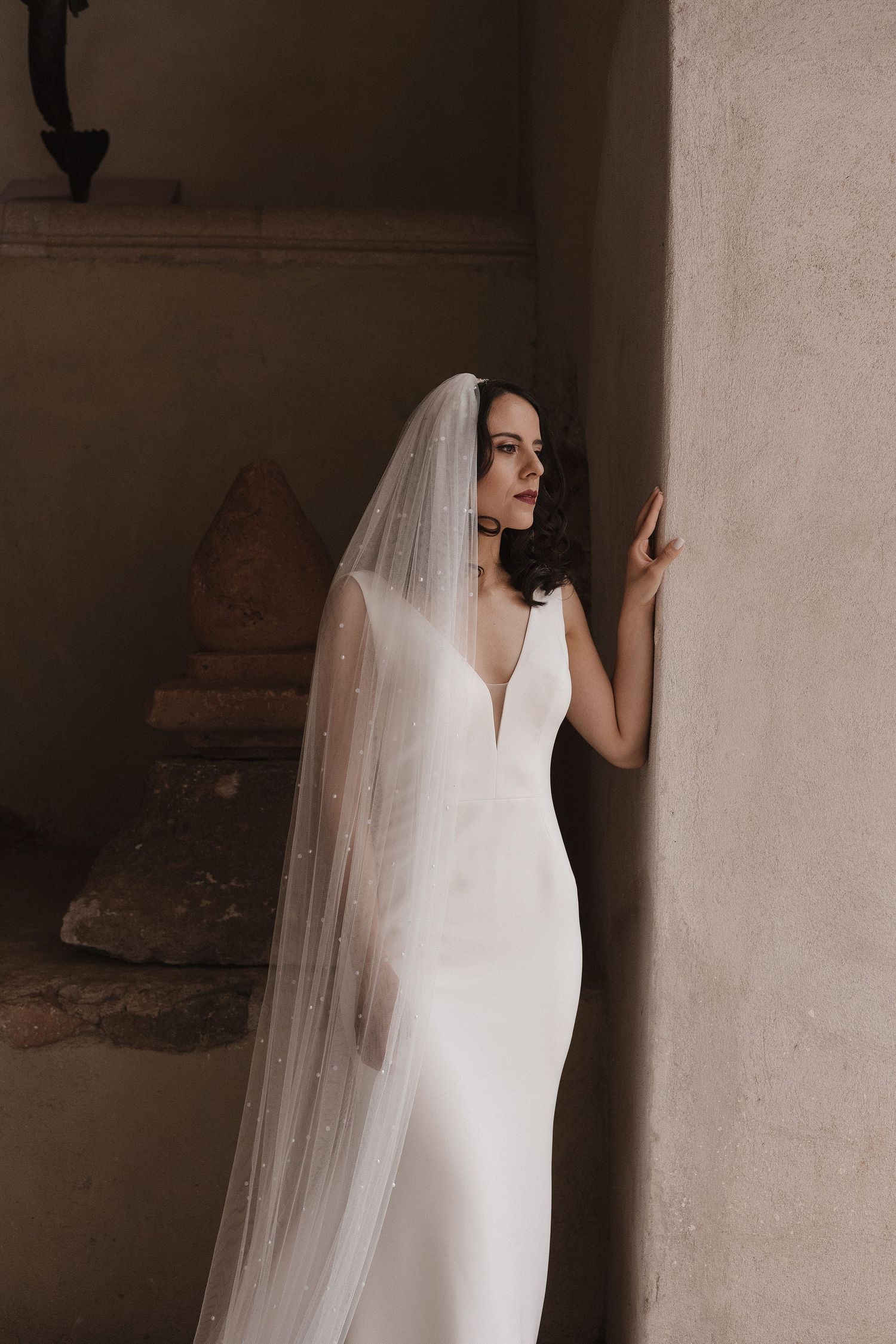 Elegant bride in a sleek white wedding dress and veil poses against a stone wall in moody lighting.