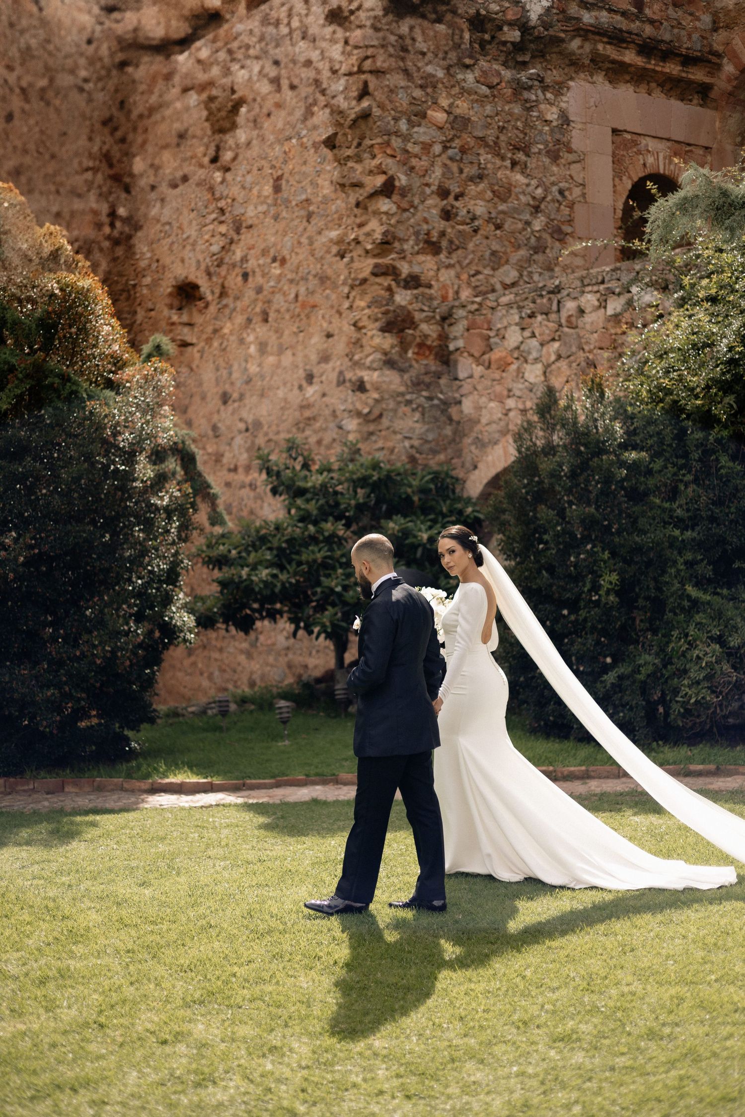 A couple in formal wedding attire sits together on a grassy lawn against a rustic stone wall backdrop.