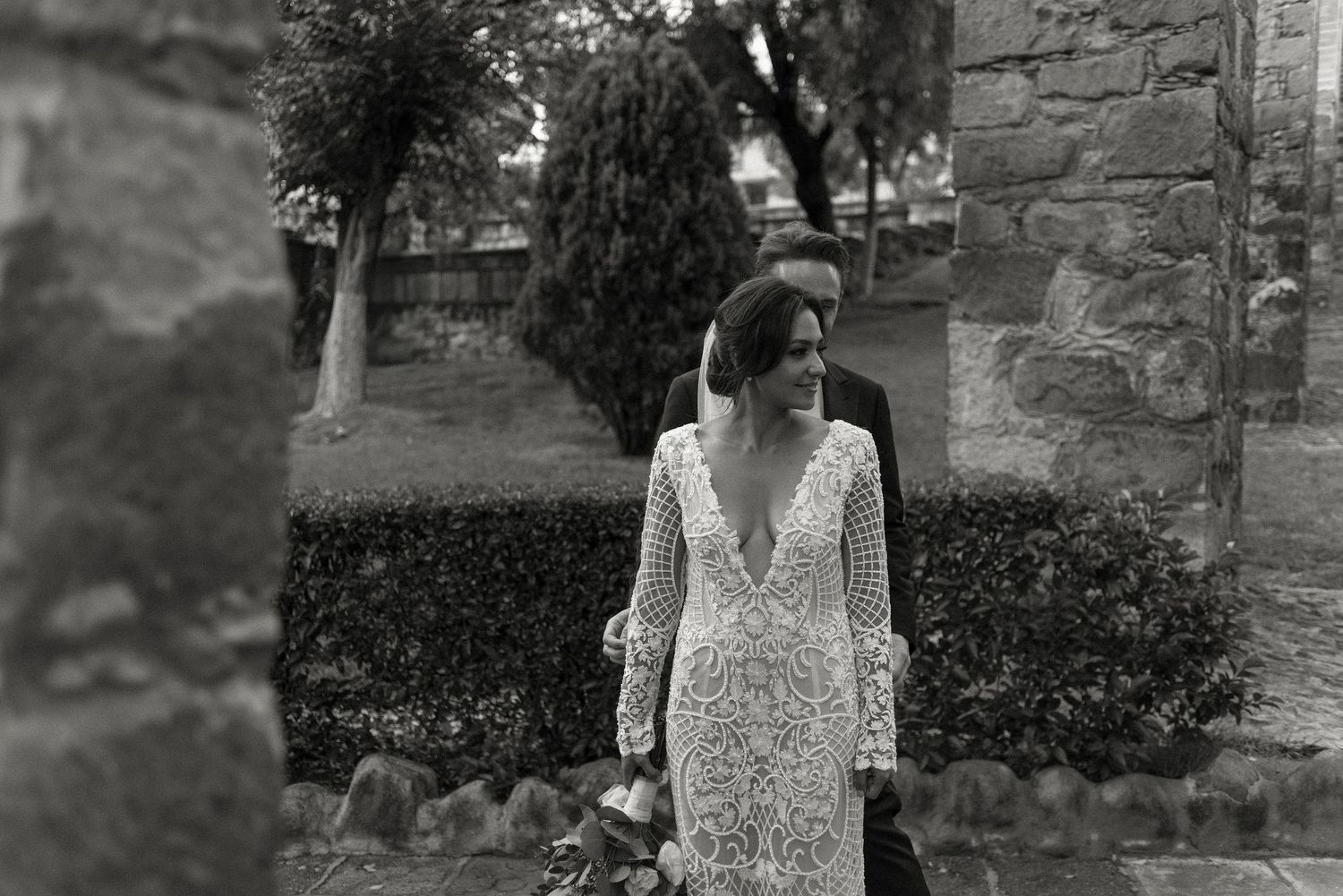 Vintage black and white photo of someone wearing a long-sleeved deep v-neck lace wedding gown in a garden setting.