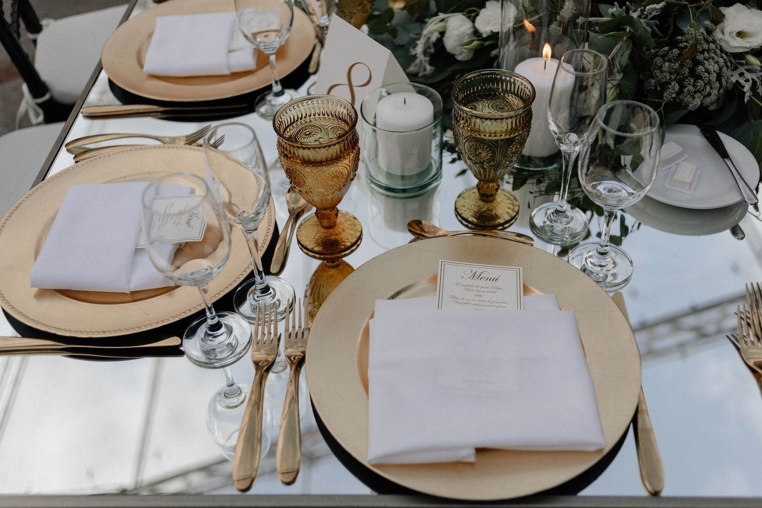 Elegant table setting with gold-rimmed plates, amber glassware, and white napkins on a white tablecloth.