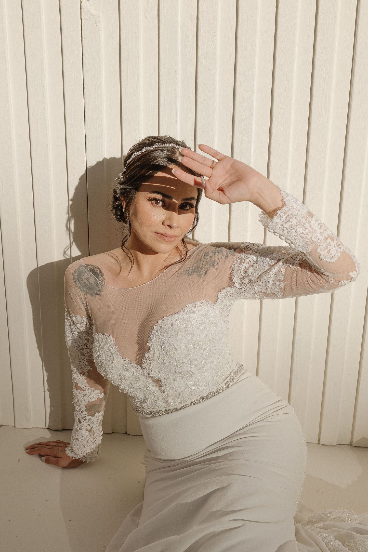 Elegant wedding dress with sheer lace sleeves and sweetheart neckline shown against white paneled wall in dramatic pose.