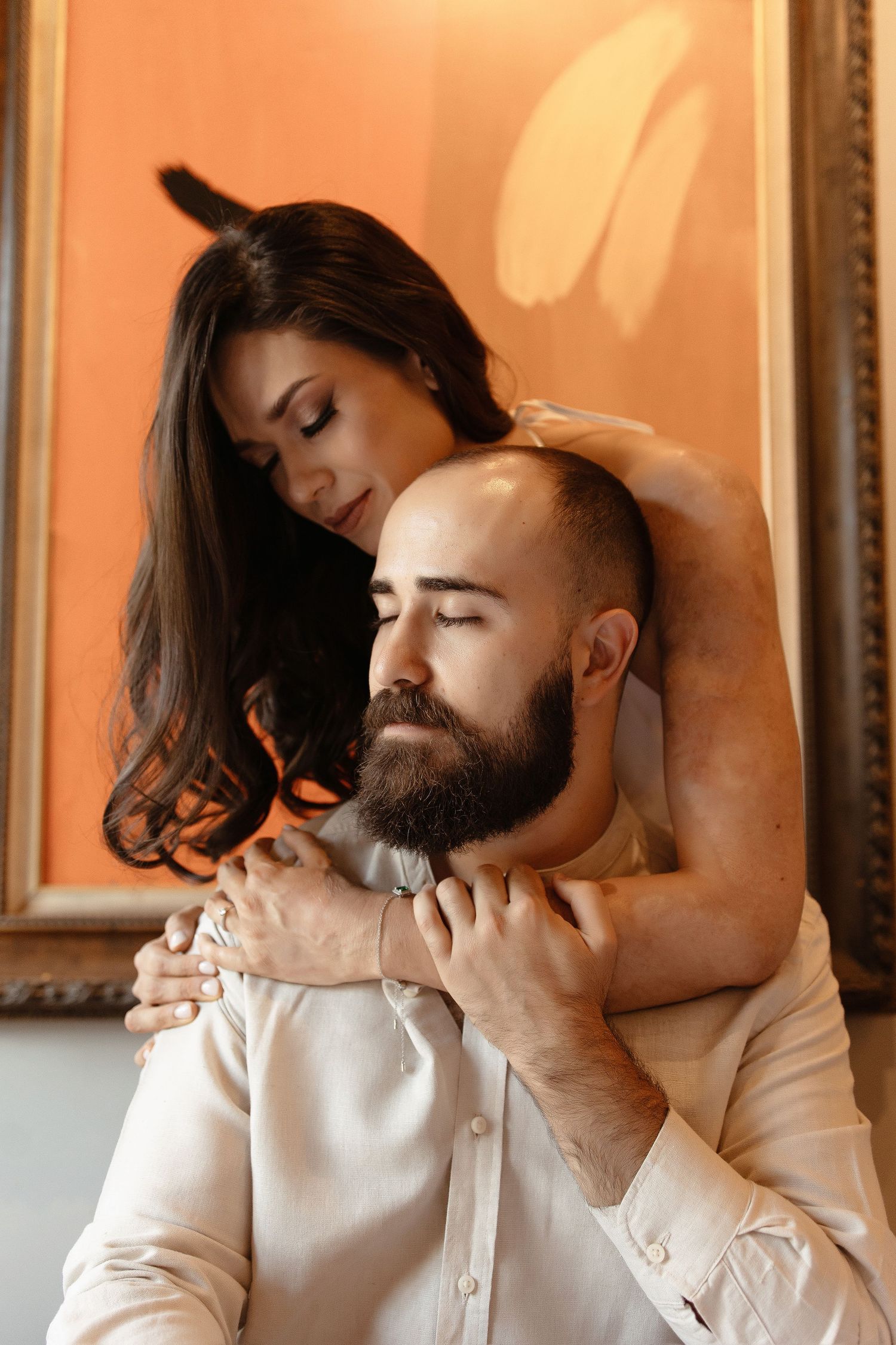 A romantic couple in white embraces intimately against a peach-colored wall with framed artwork.