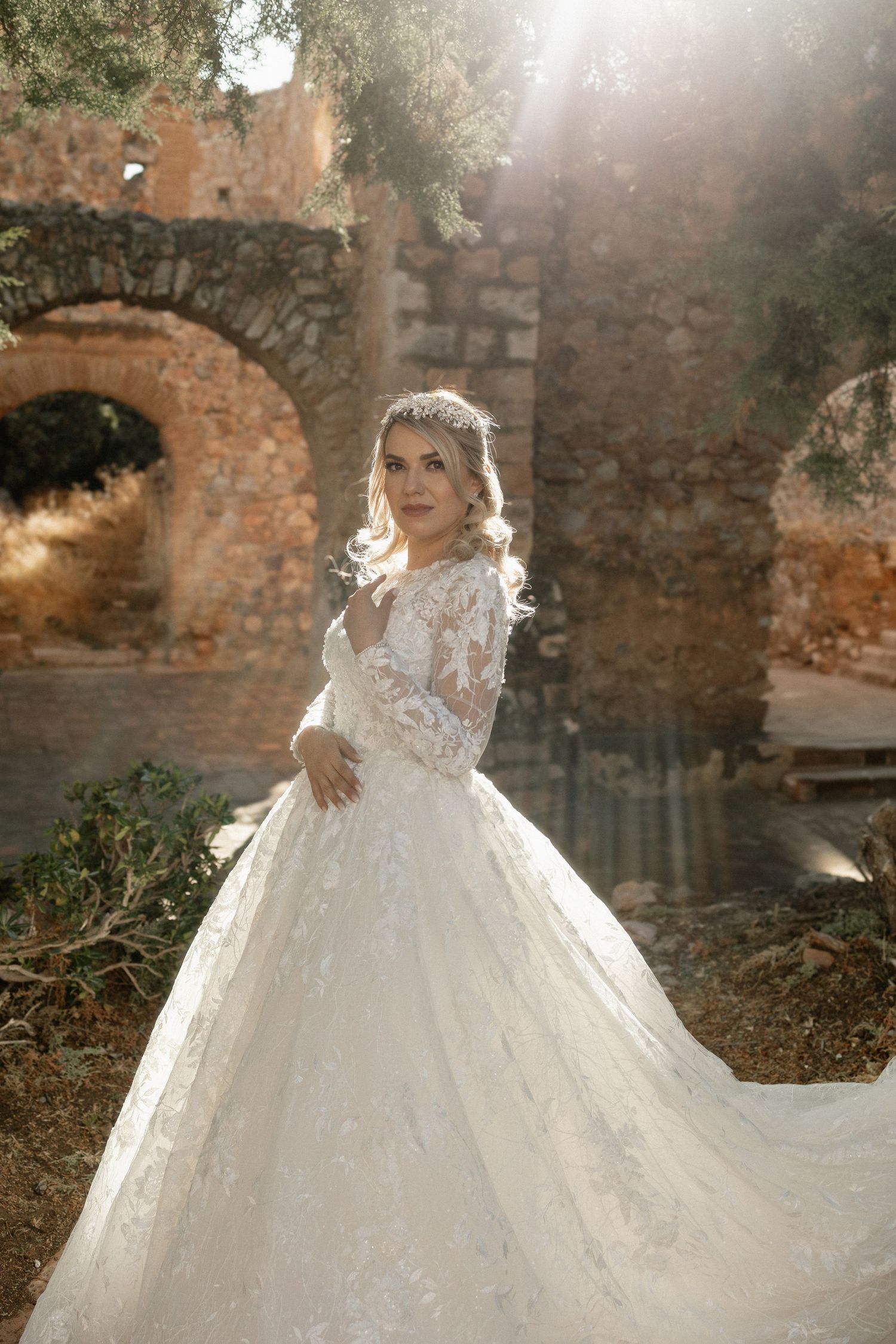 Elegant white lace wedding gown with long sleeves and full skirt photographed against ancient stone ruins at sunset.