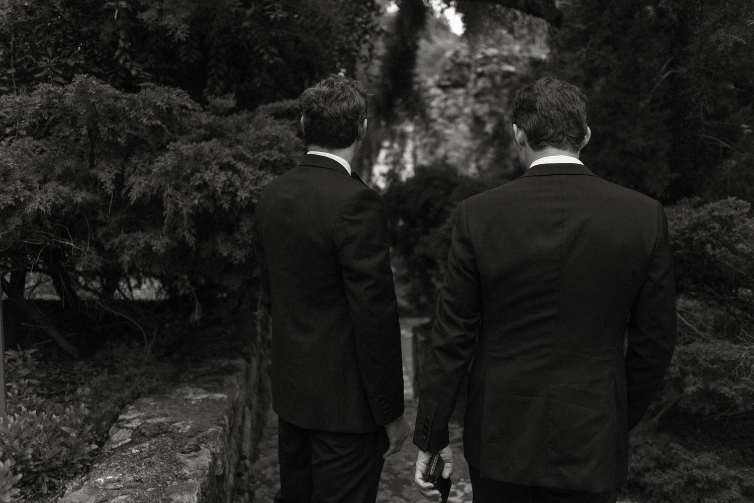 Two people in black suits stand with their backs to the camera in an outdoor garden setting.