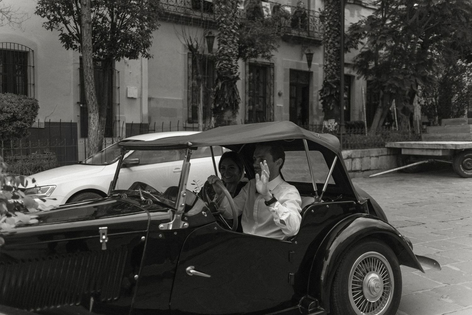 A vintage convertible automobile parked on a peaceful tree-lined street in a black and white photograph.