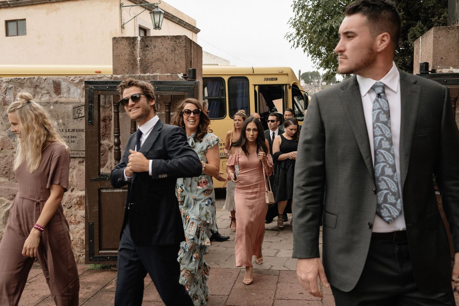 Wedding guests in formal attire walk past a yellow school bus outside a venue on a sunny day.