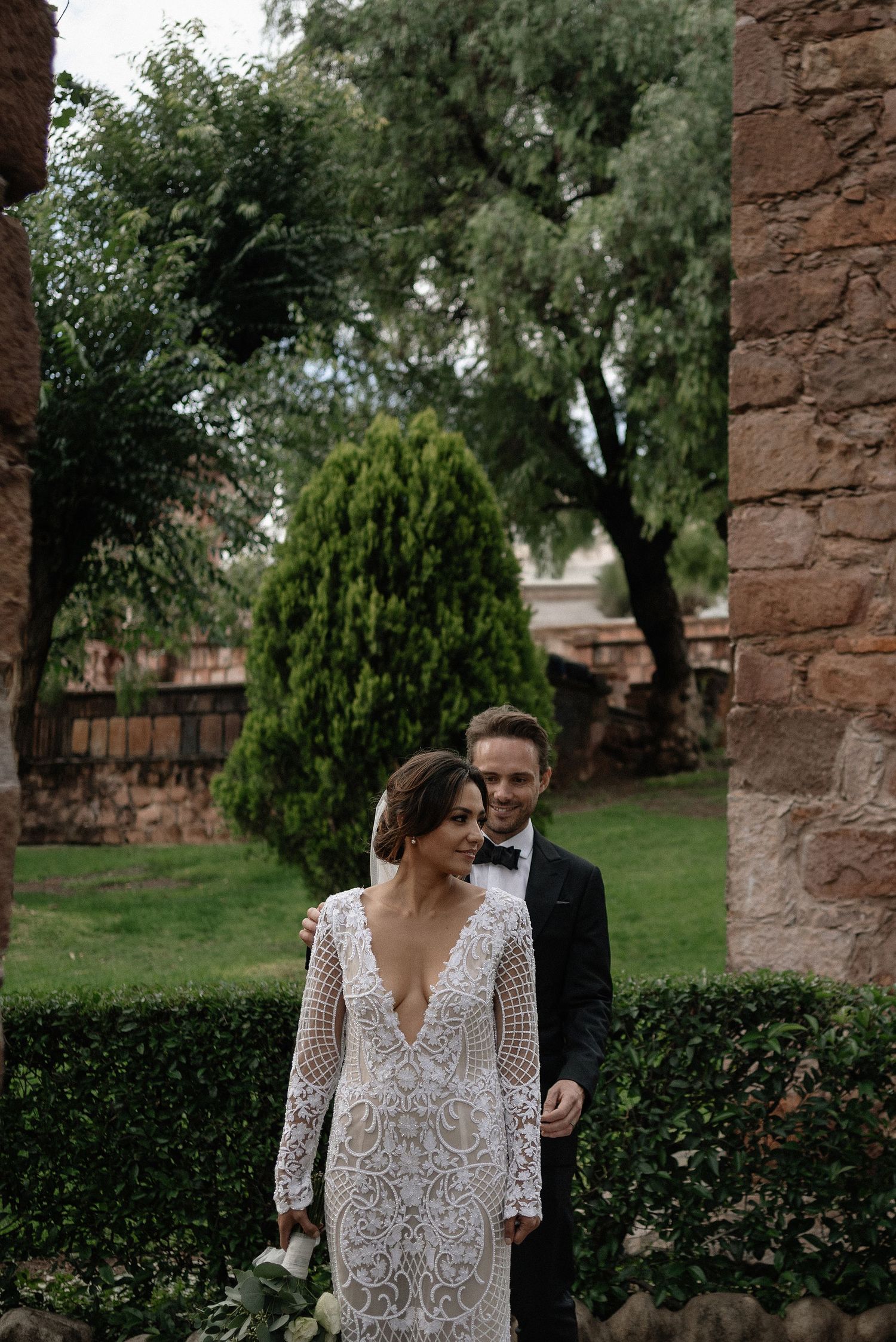 A couple in their wedding attire stand near a stone wall with lush greenery and trees in the garden background.