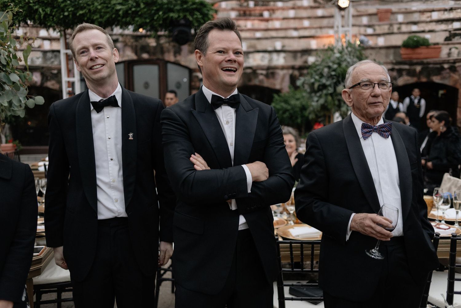 Three men in black tuxedos and bow ties stand together at an outdoor event reception.