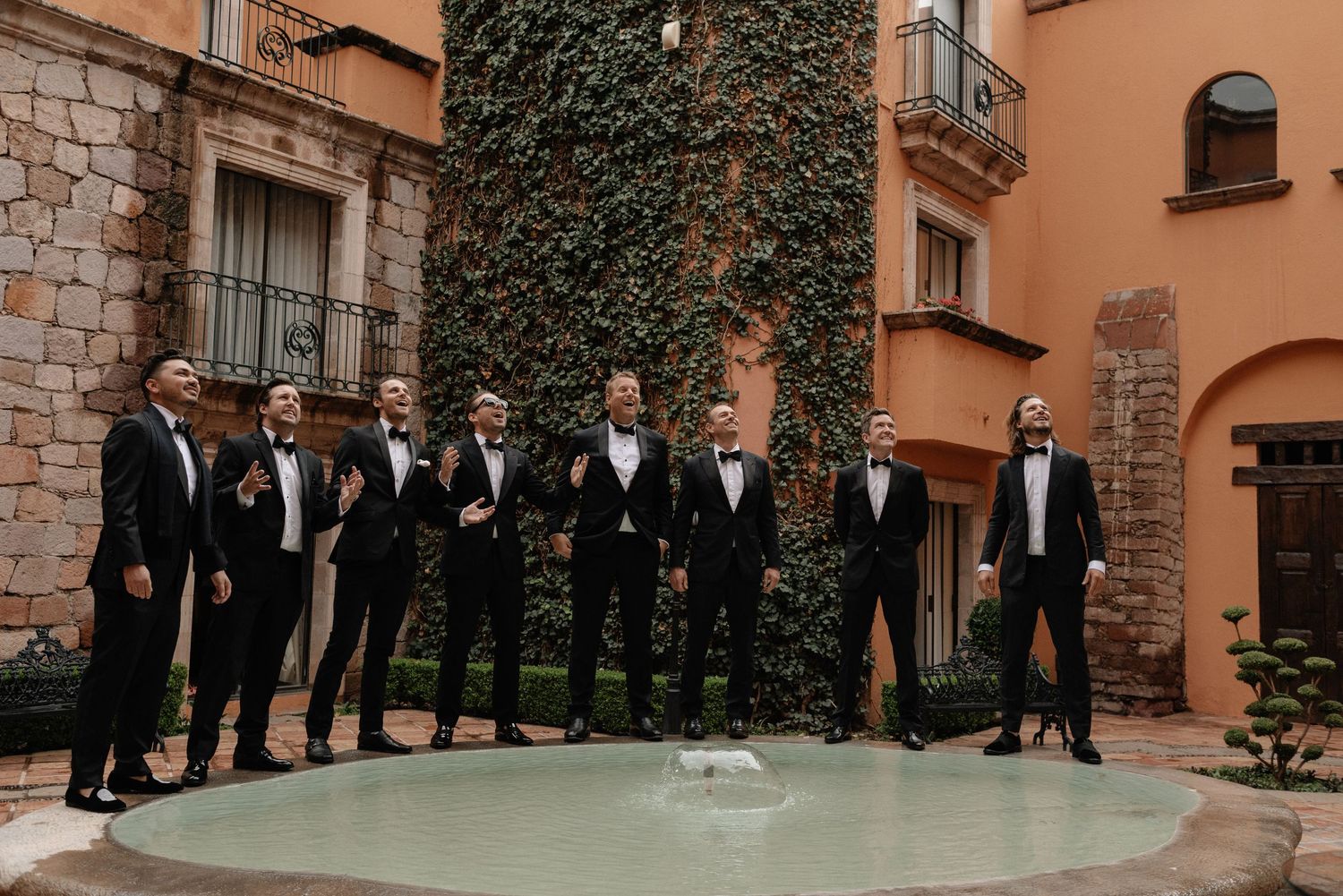 A group wearing black tuxedos poses together against an ivy-covered wall at an elegant venue.