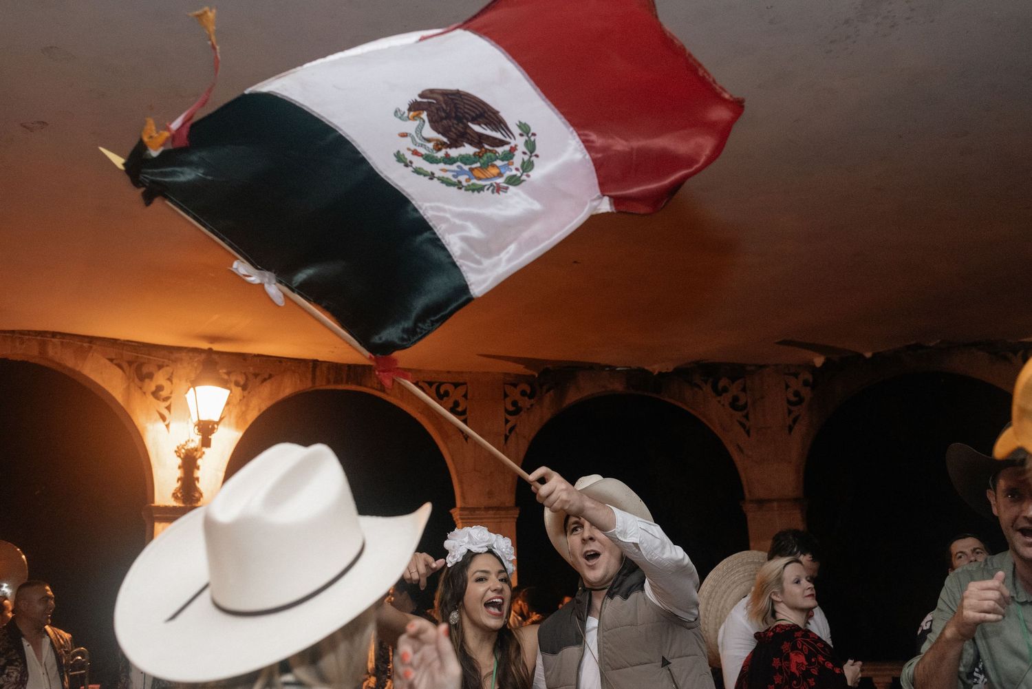 Mexican flag waving above a festive celebration at night with people wearing cowboy hats and sombreros.