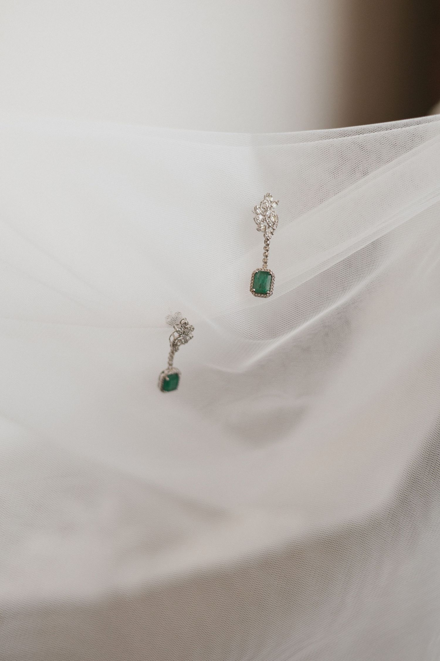 Small emerald and diamond drop earrings displayed on delicate white tulle fabric in soft lighting.