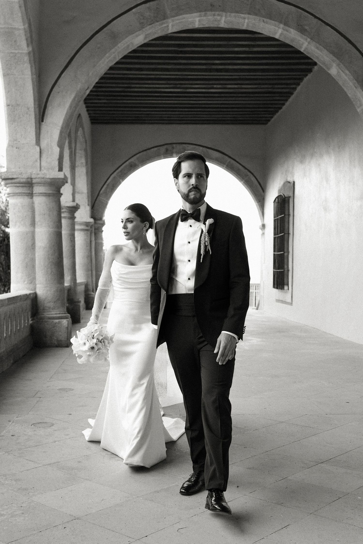 Black and white photo of elegant bride and groom walking under stone arches at a classic wedding venue.