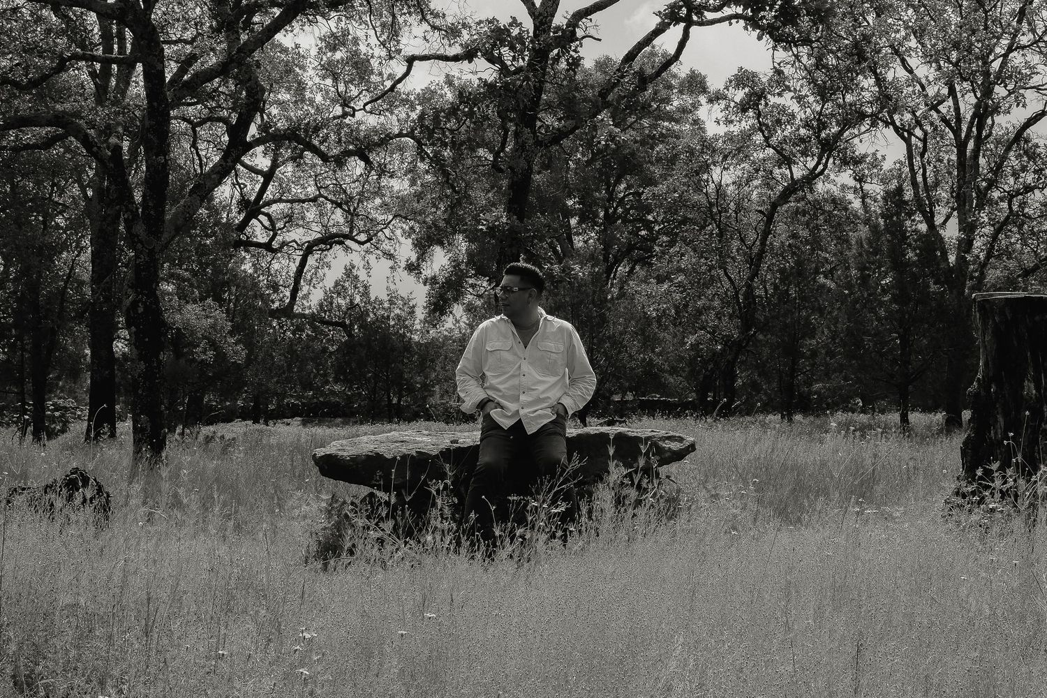 Black and white photo of someone seated on wooden bench in grassy field with trees.